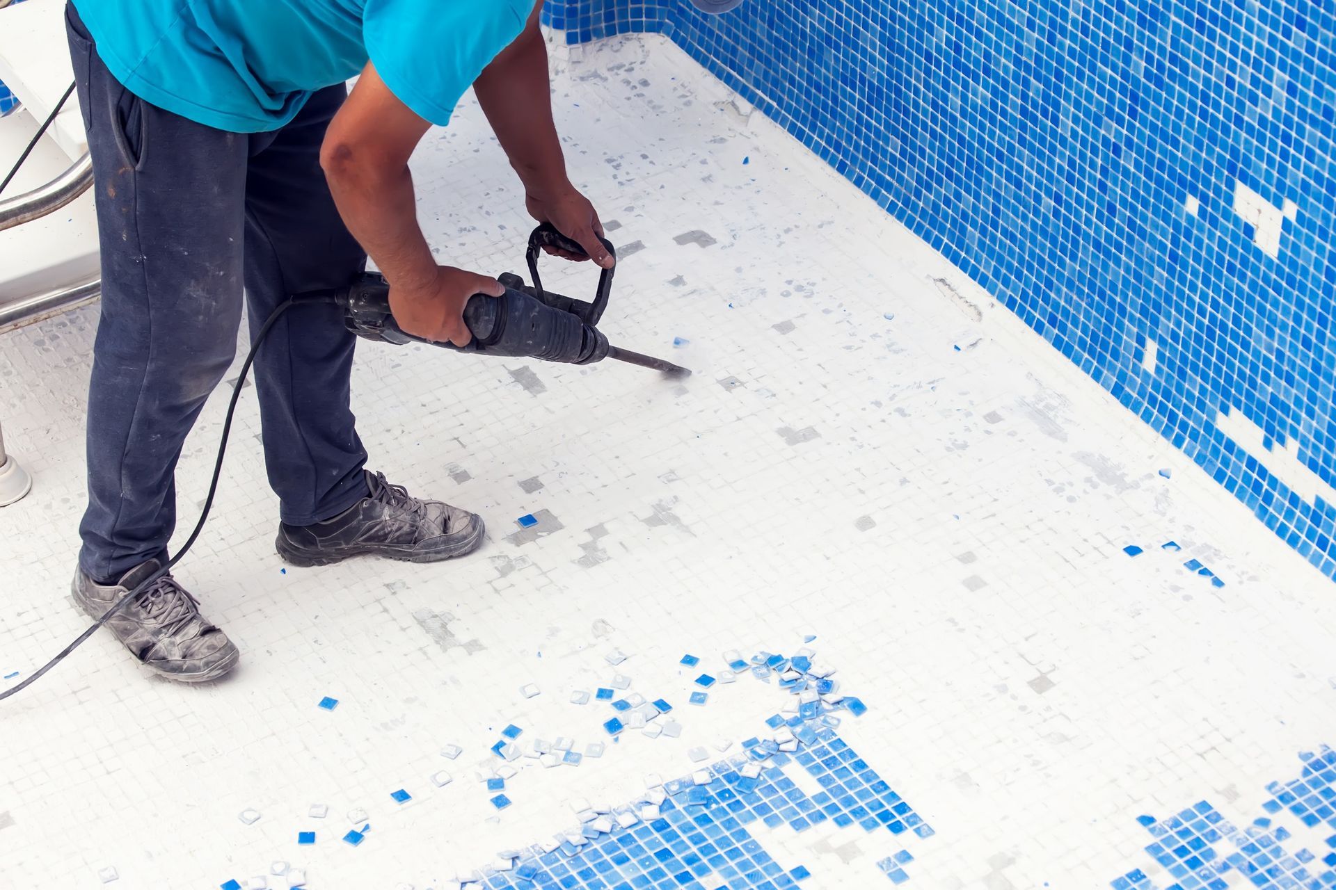 A worker using a power tool to remove old tiles from the floor and walls of a blue-tiled swimming pool.