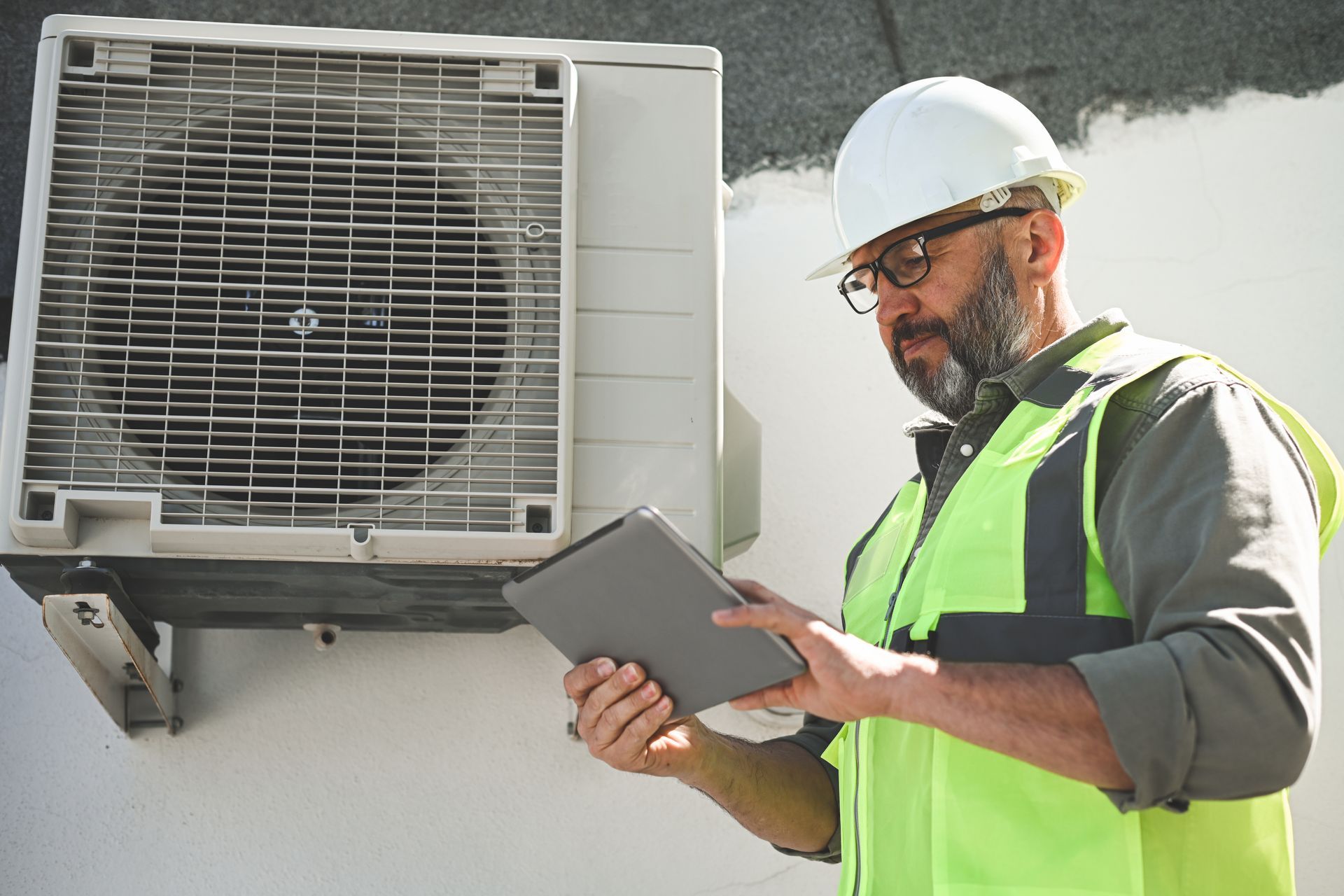 A man in a hard hat and safety vest is looking at a tablet in front of an air conditioner.