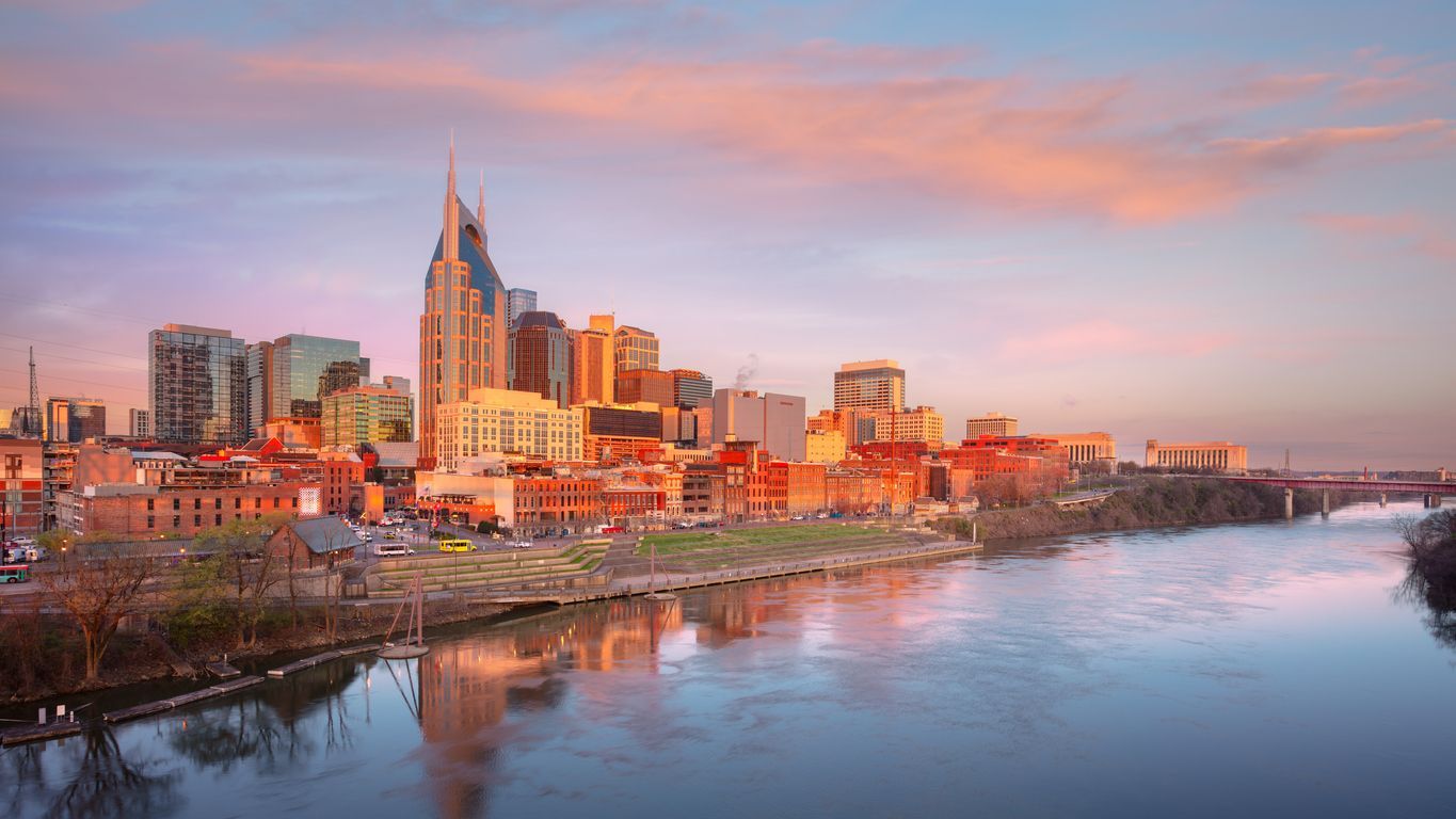 Nashville skyline at sunset, reflected in the Cumberland River. Buildings are lit with golden light and pink clouds.