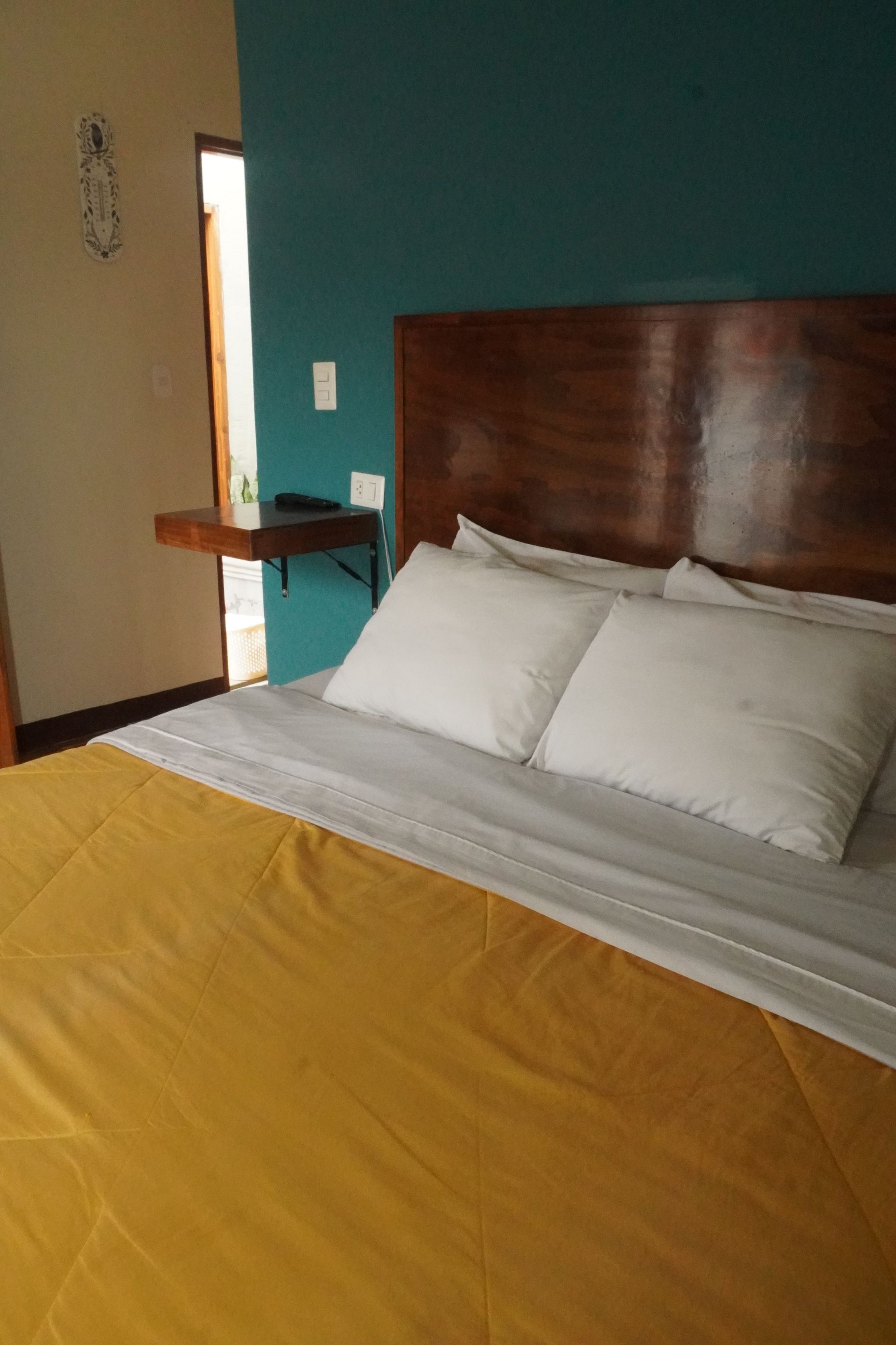 Bedroom with a bed covered in a yellow blanket, white pillows, and a wooden headboard. A teal wall is behind the bed.