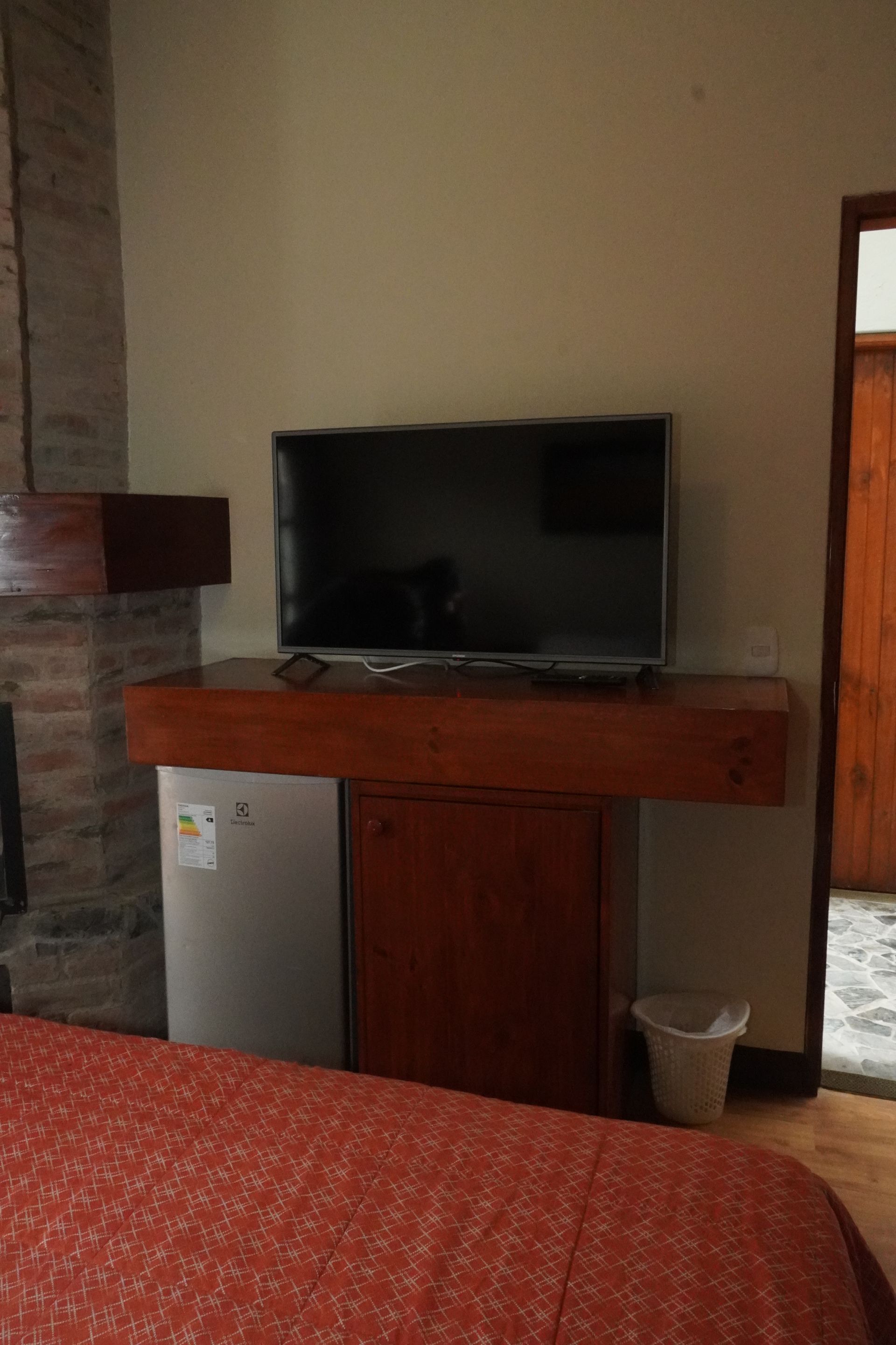 A hotel room with a TV, minibar, and a view of a doorway; the bed has a red patterned cover.
