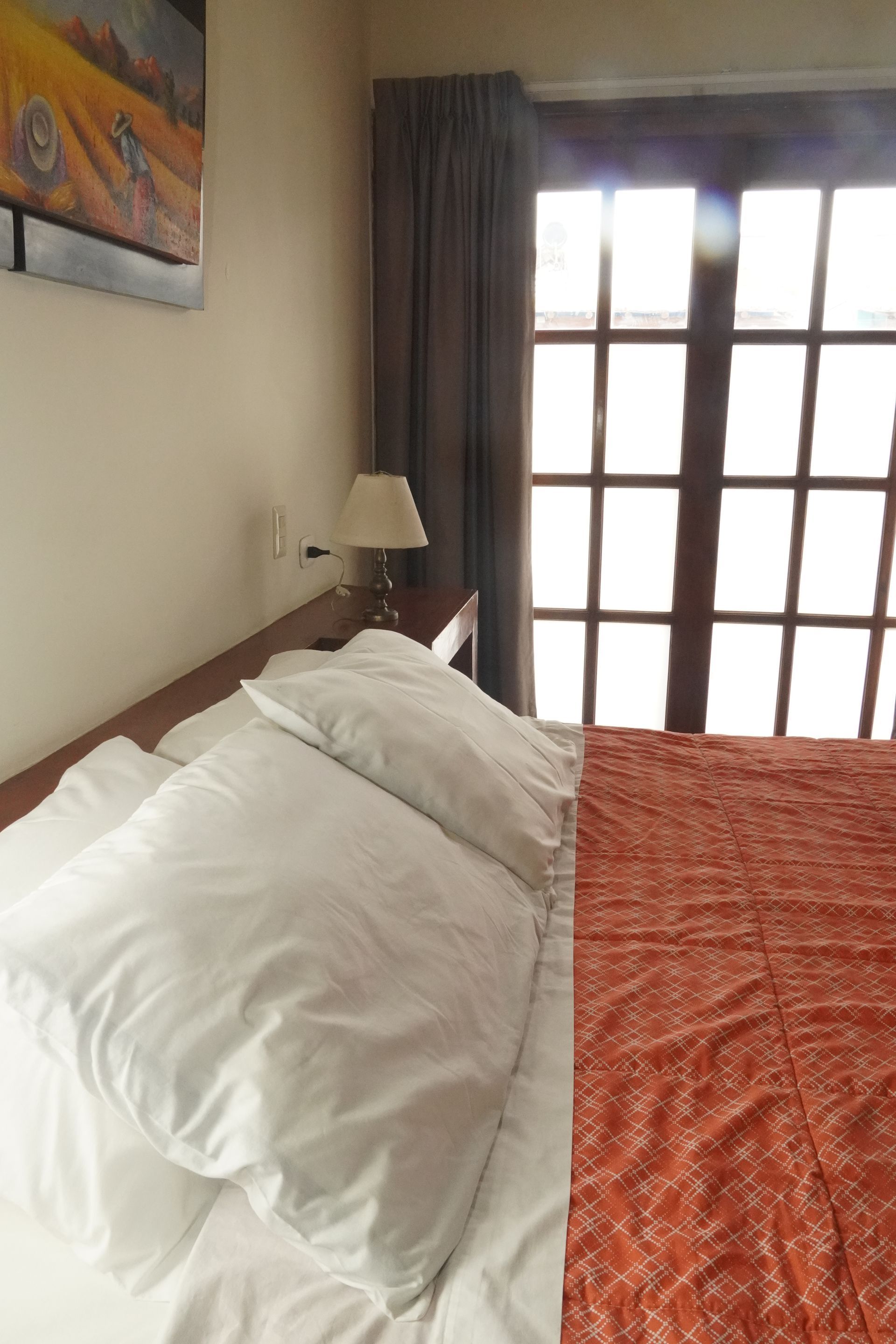 Bed with white pillows and orange bedspread, next to a window with sunlight.