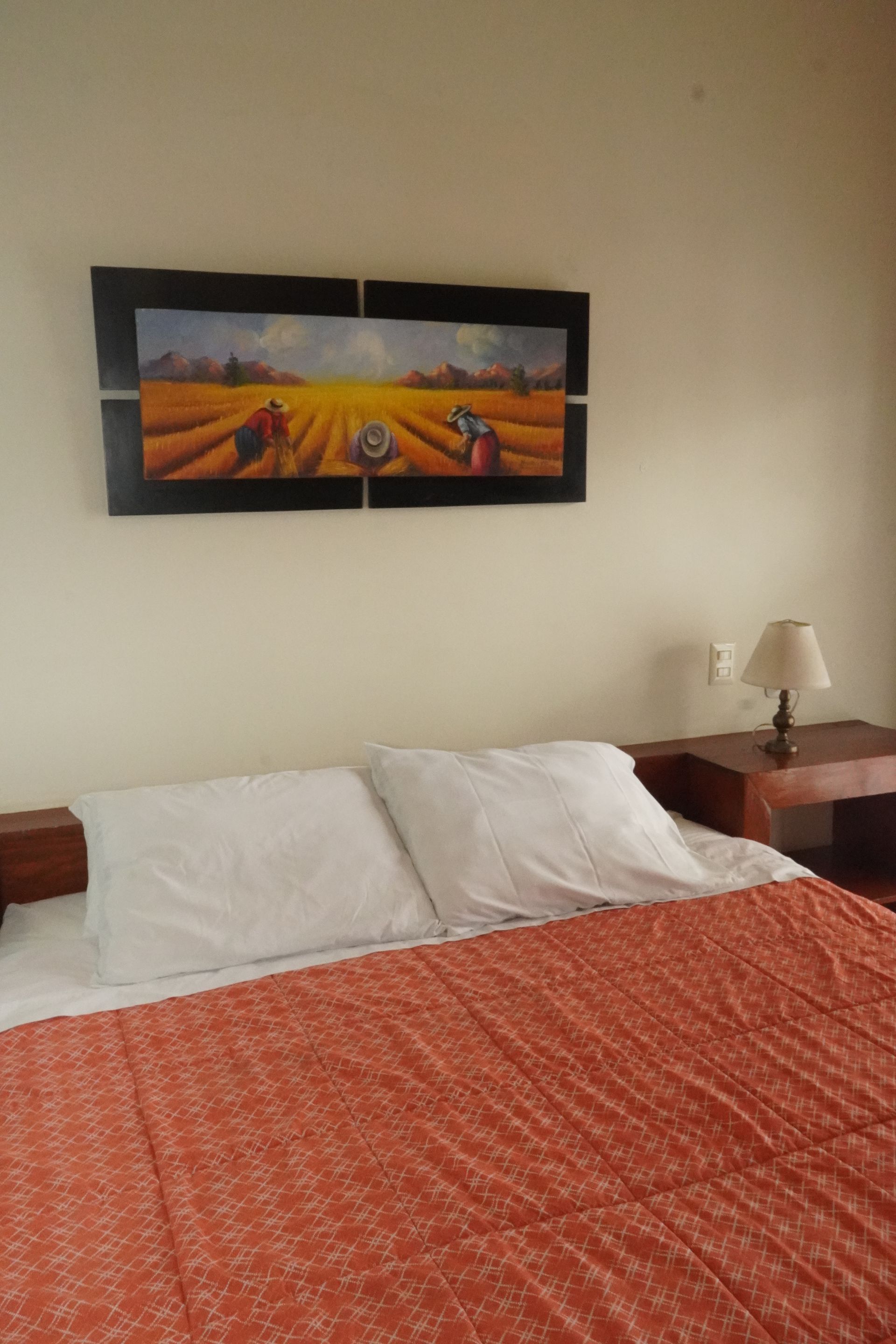 Bed with orange quilt, white pillows, bedside table with lamp, and landscape painting on wall.