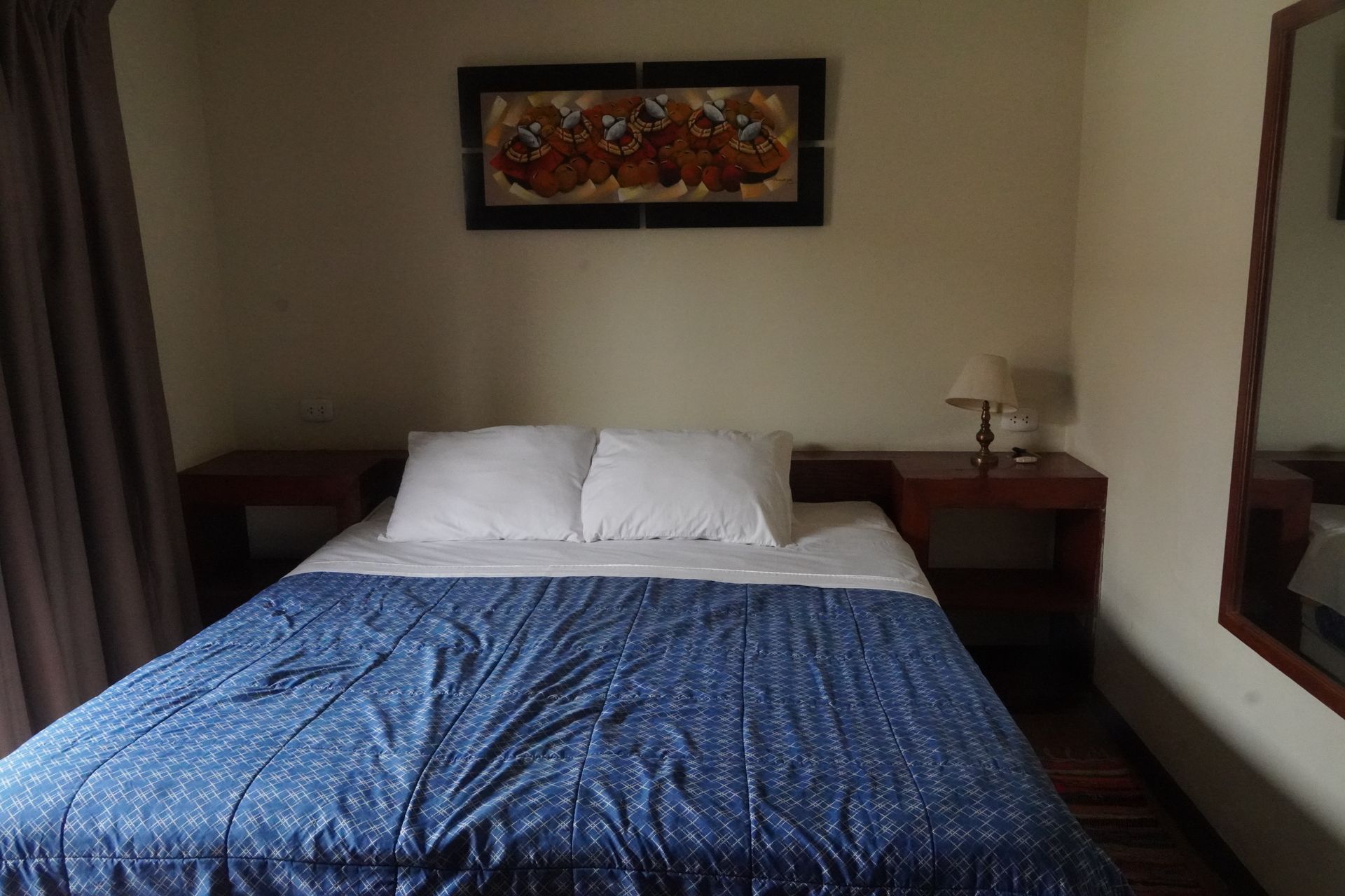 Bedroom with bed, nightstands, lamp, artwork, and mirror. Blue comforter and white pillows.