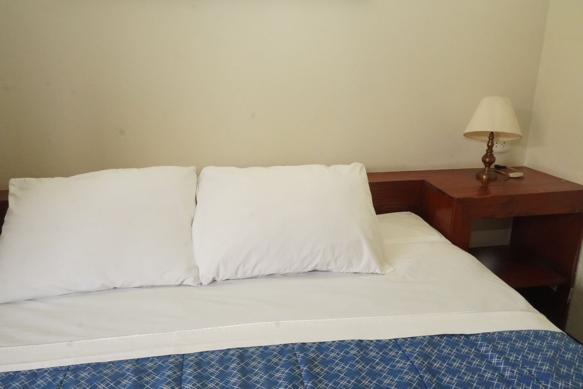 Bed with white pillows and blue blanket, beside a nightstand with a lamp.
