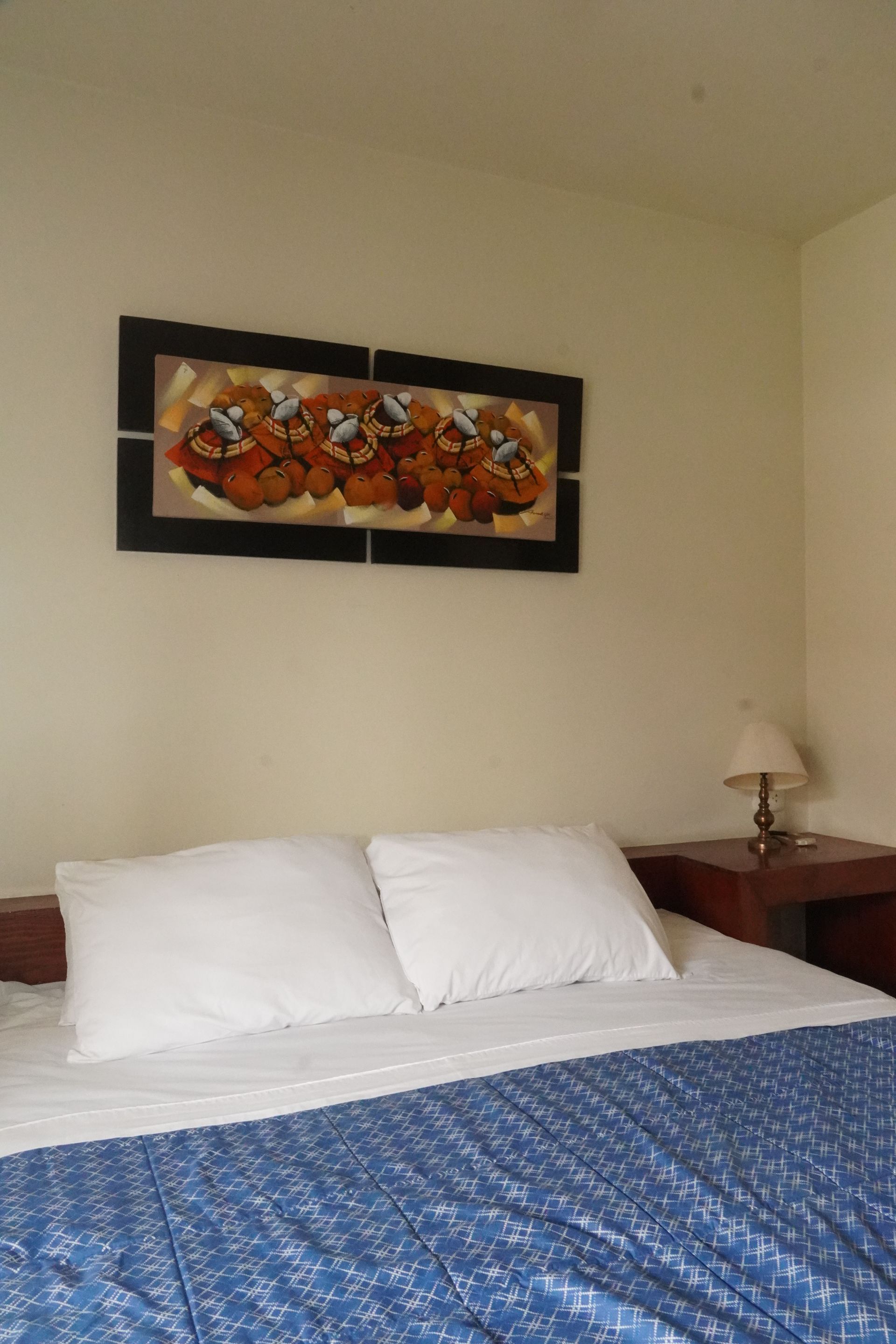 Bedroom with bed, pillows, blue quilt, painting, nightstand, and lamp.