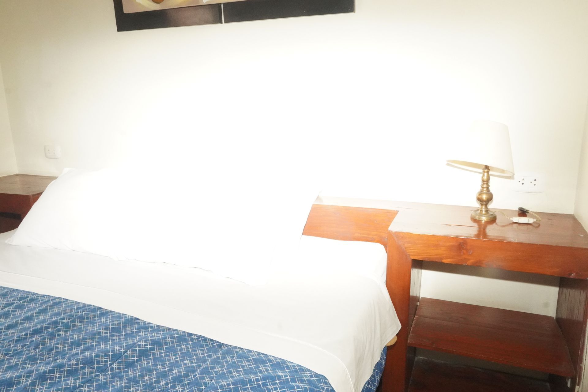 Bedroom with bed, white sheets, blue blanket, wooden nightstand, lamp, white wall.