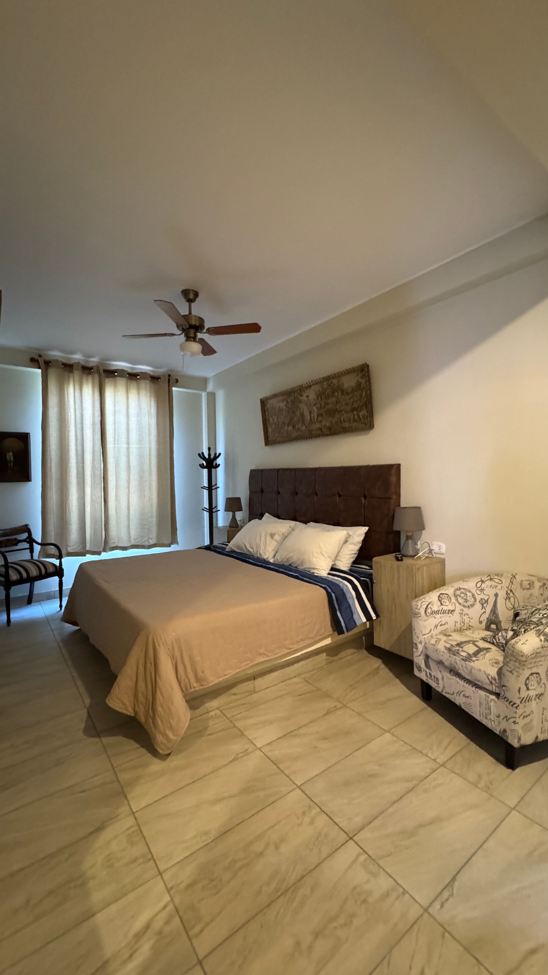Bedroom with bed, patterned armchair, and beige tiled floor. Sunlight streams in from a window with curtains.