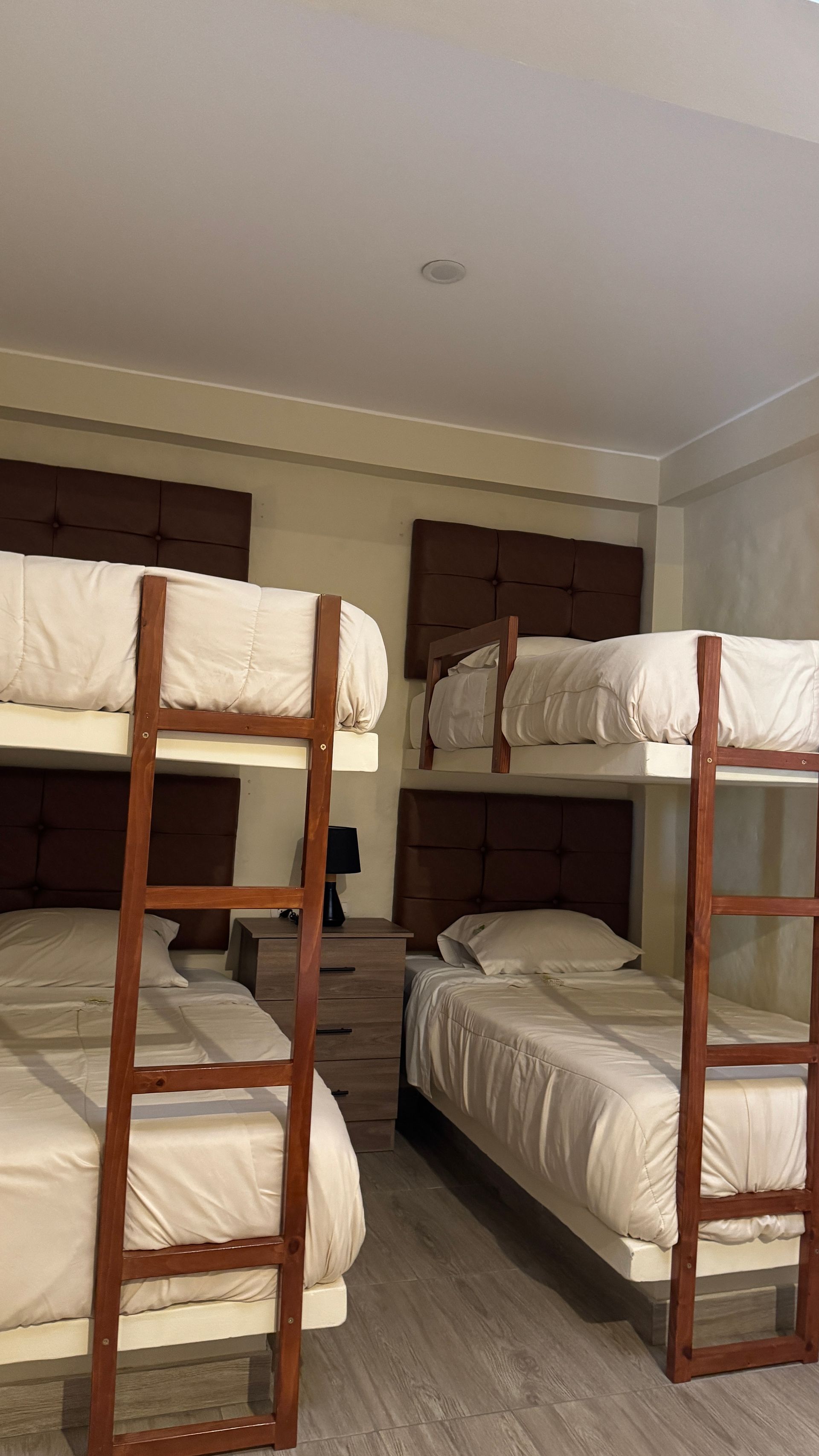 Bunk beds in a room. Three sets of beds with white bedding, brown ladders, and headboards. A nightstand is between two beds.