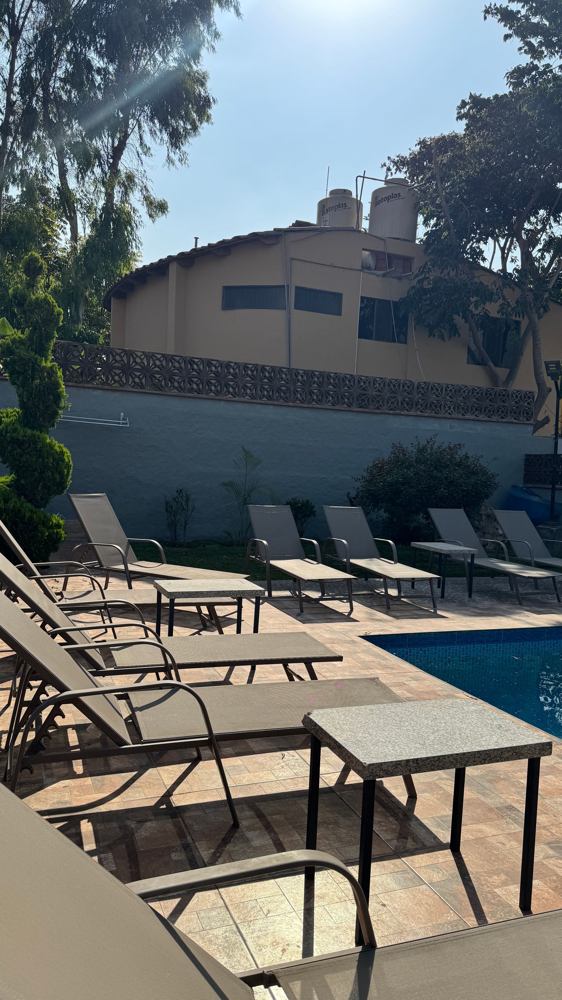 Poolside scene with lounge chairs, blue water, building, trees, and bright sky.