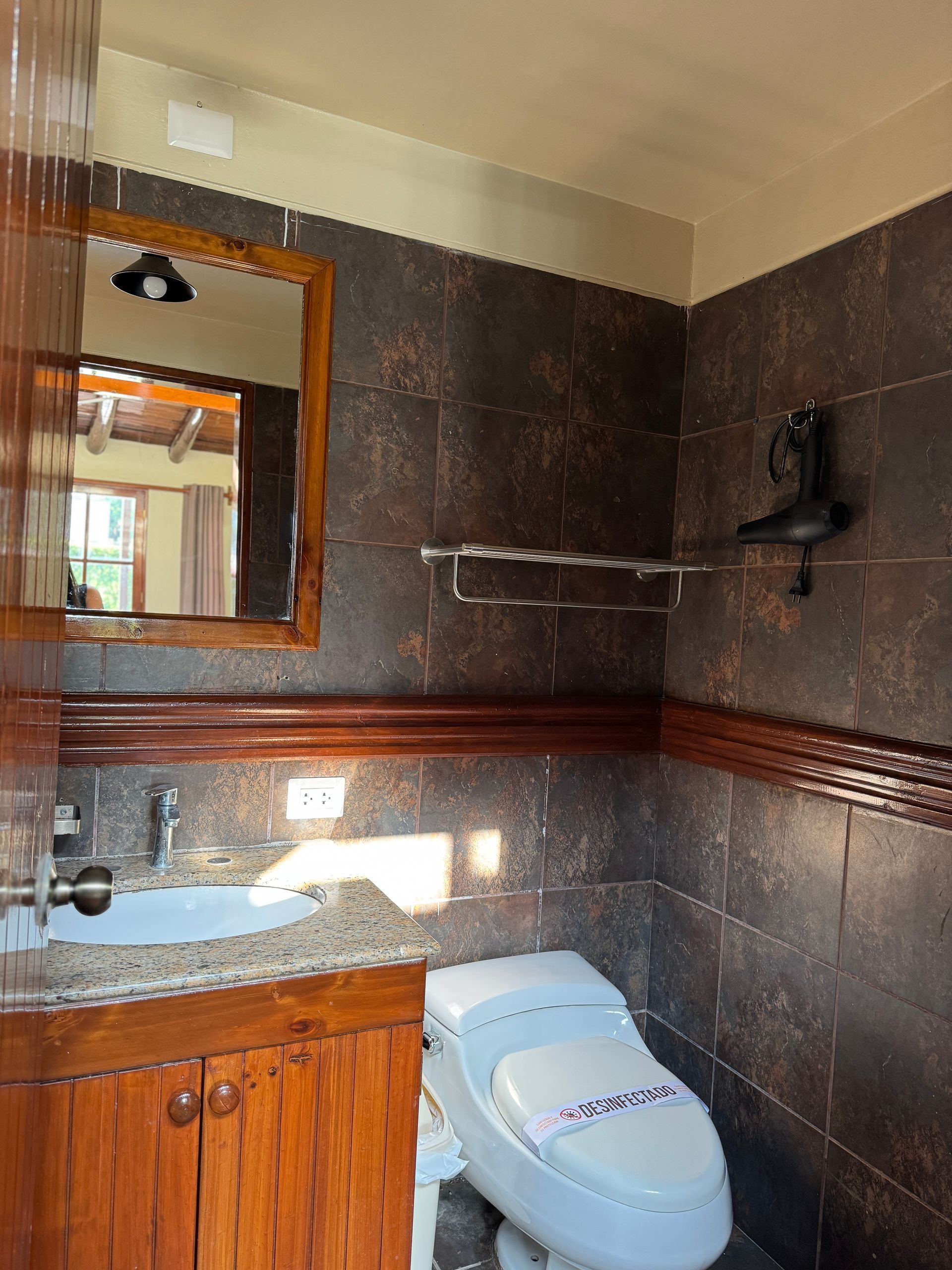 Bathroom with brown tile walls, wooden vanity, and toilet. Mirror and towel rack visible.