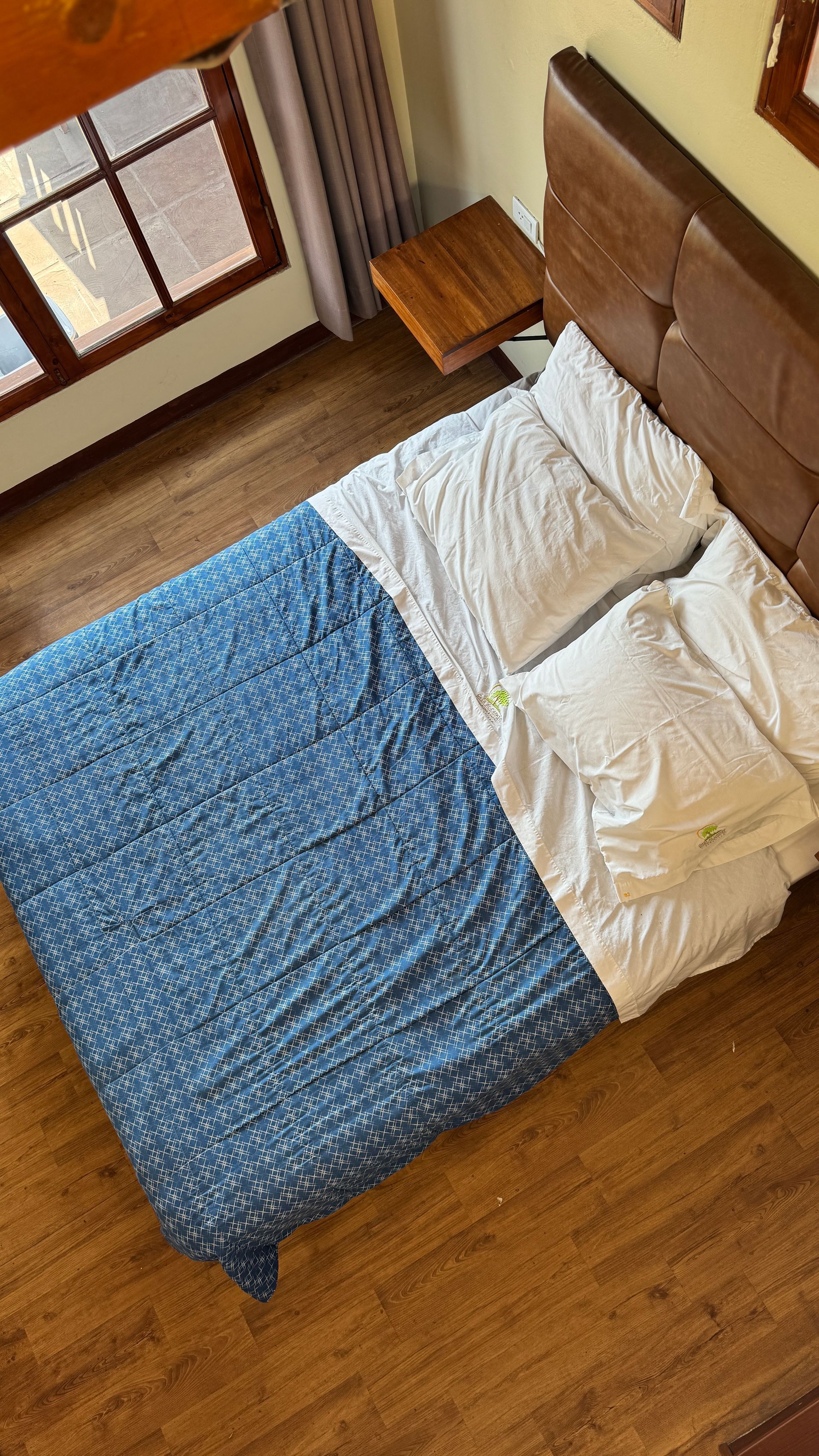 Bed with blue patterned blanket, white sheets, wooden headboard, and bedside table next to a window.