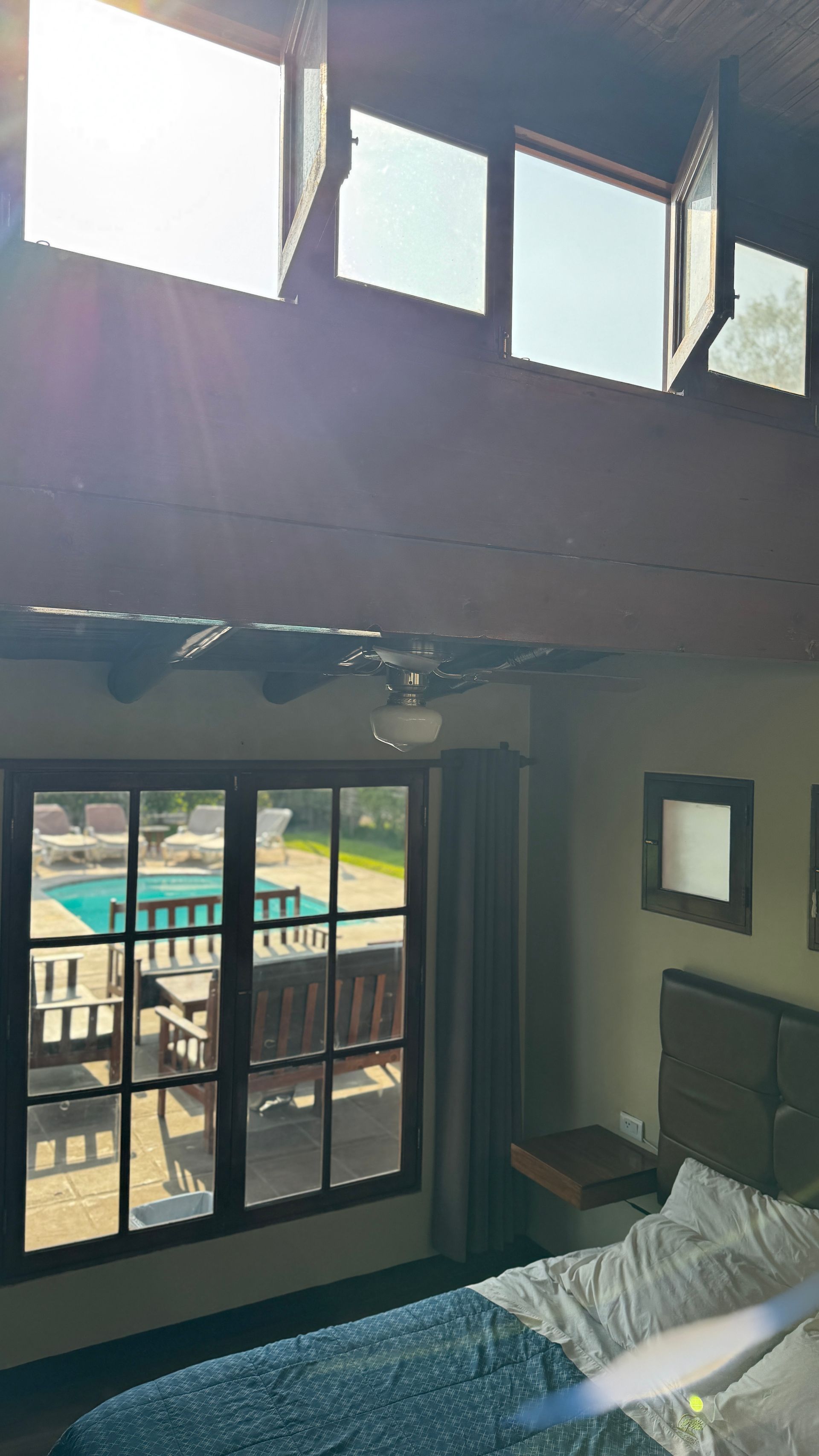 Bedroom interior with large window overlooking a pool, wood beams, and a row of small windows above.