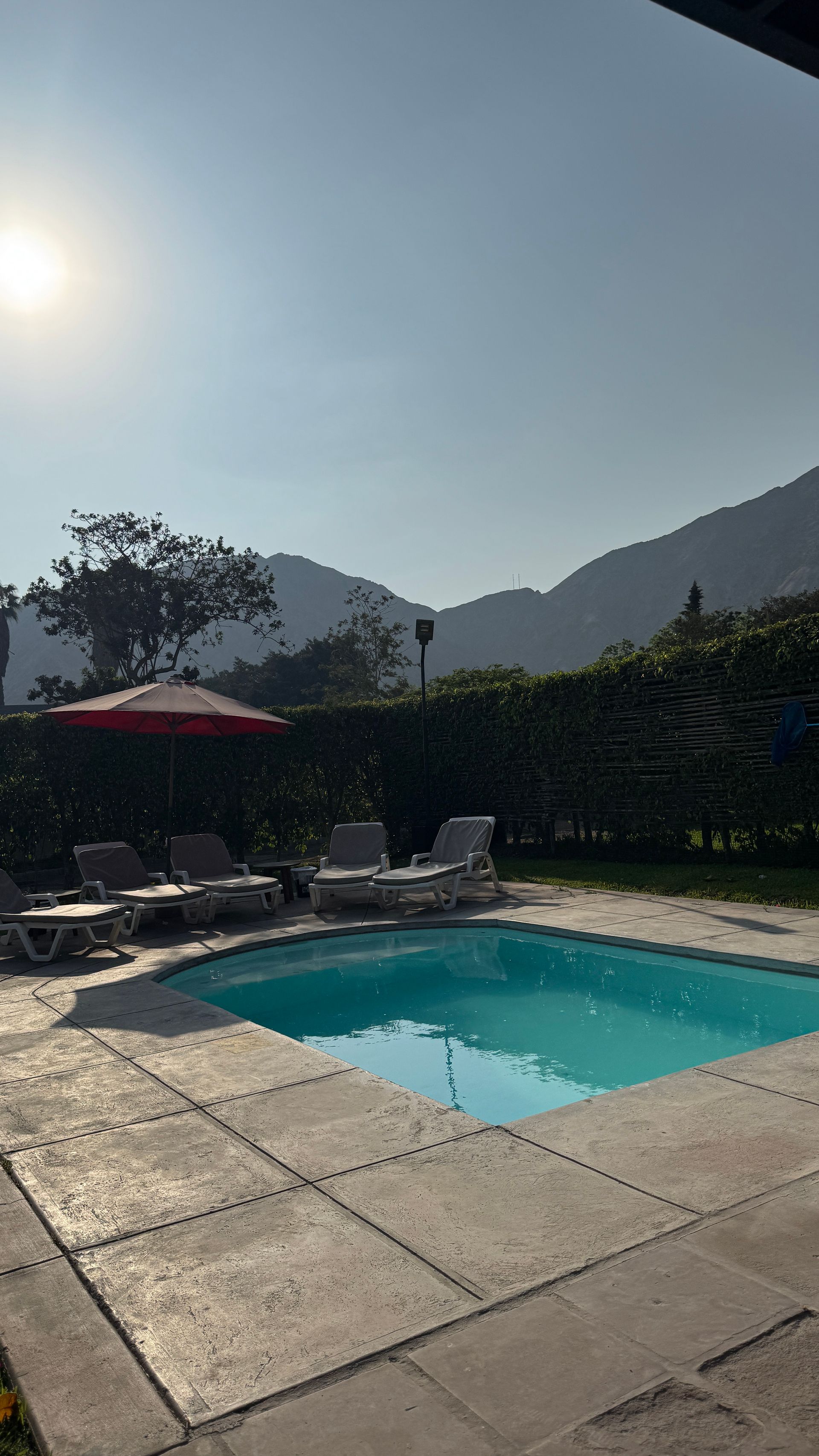 Pool with lounge chairs and umbrella, mountains in the background under a sunny sky.