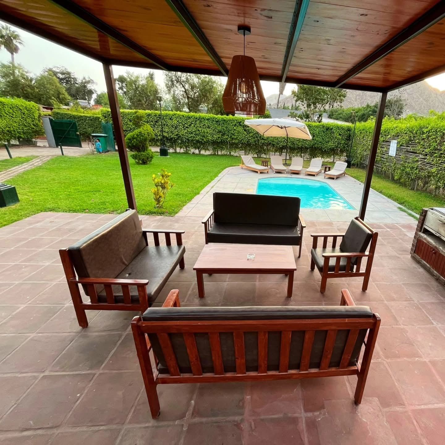 Patio with wood furniture, pool, and green yard under a shaded pergola.
