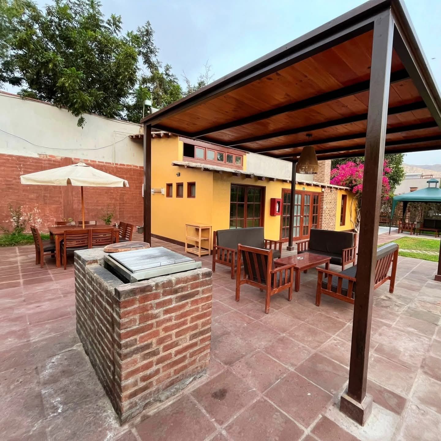 Patio with brick barbecue, wooden furniture, and yellow house under a wood-covered pergola.