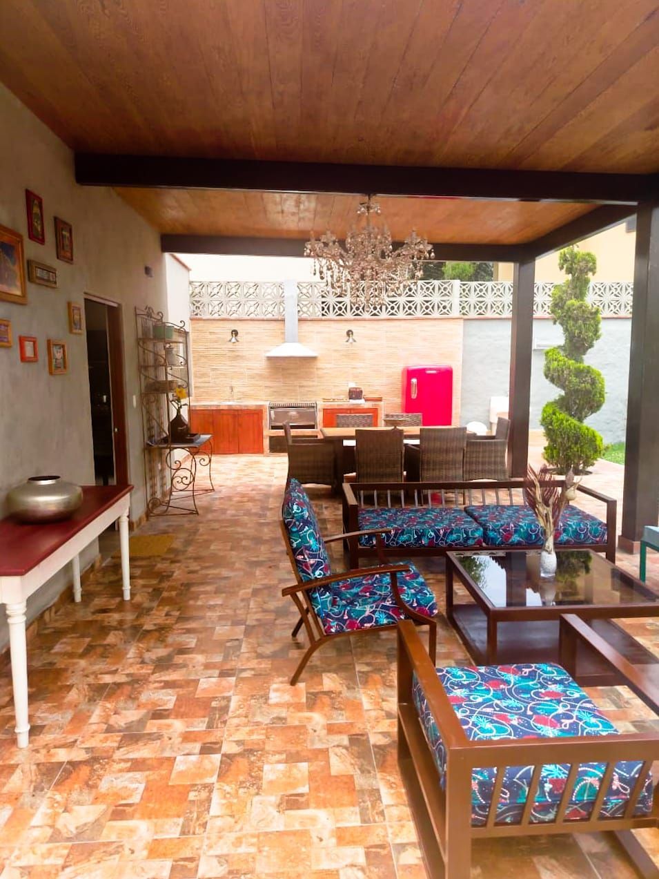Outdoor patio with seating, dining area, and kitchen. Wooden pergola, tiled floor, and chandelier.