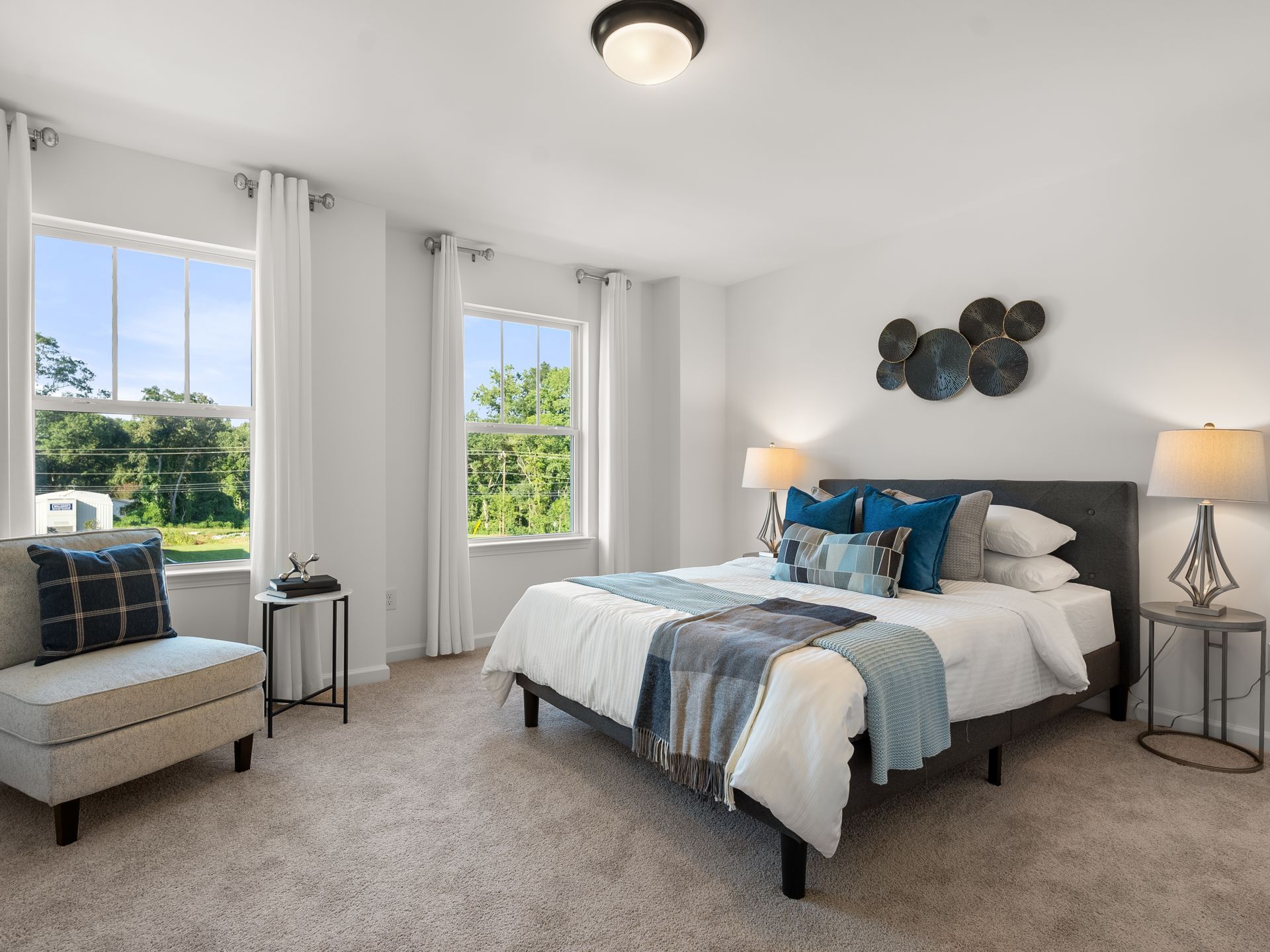 A bedroom with a bed , chair , lamps and two windows.