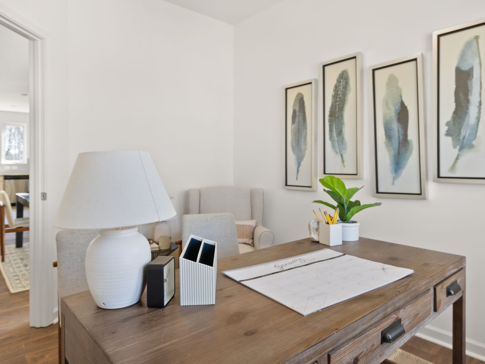 A home office with a desk , chair , lamp and paintings on the wall.