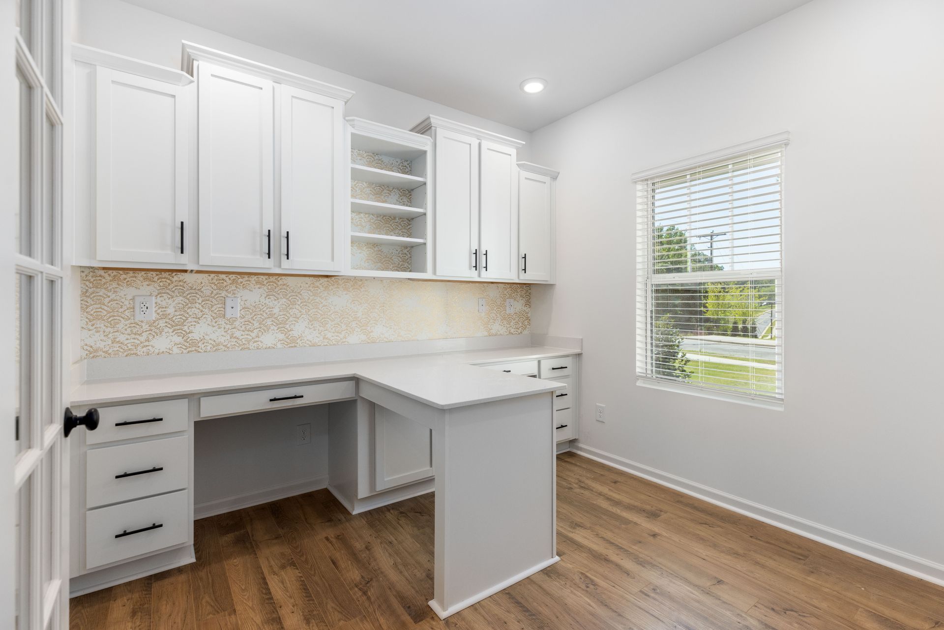 A kitchen with white cabinets , hardwood floors , a desk and a window.