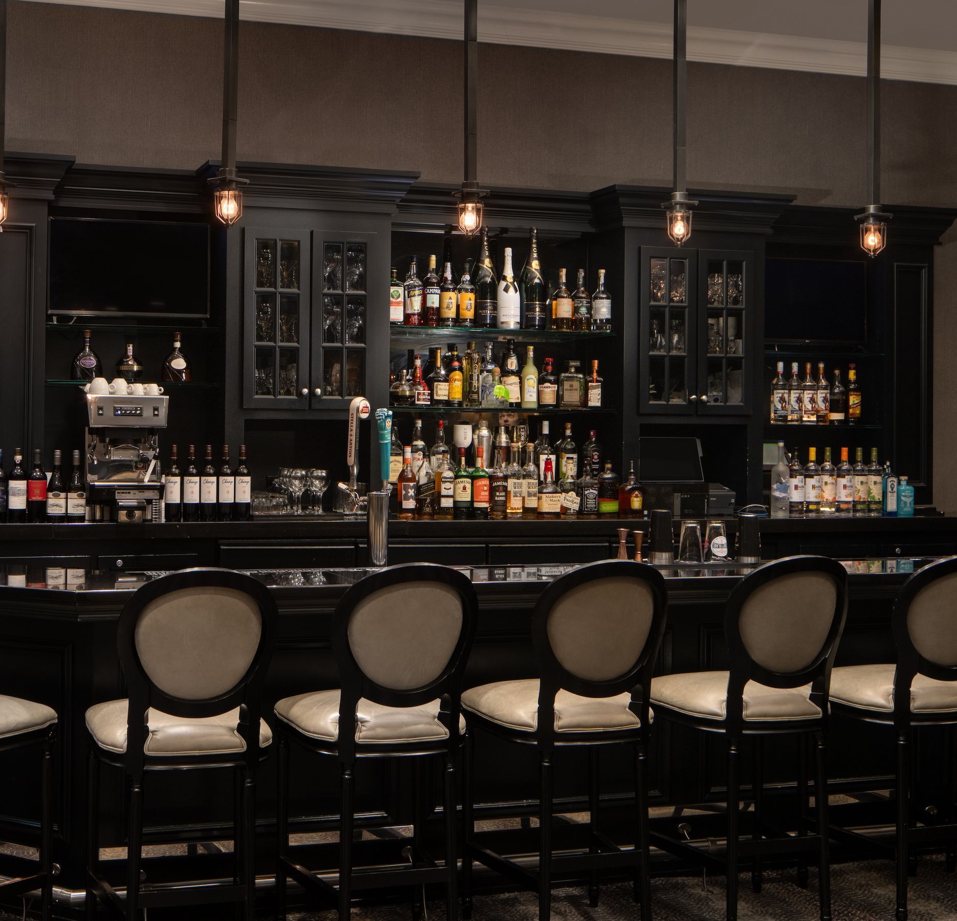 Bar with black cabinetry, liquor bottles, and high-backed chairs. Overhead lights illuminate the space.