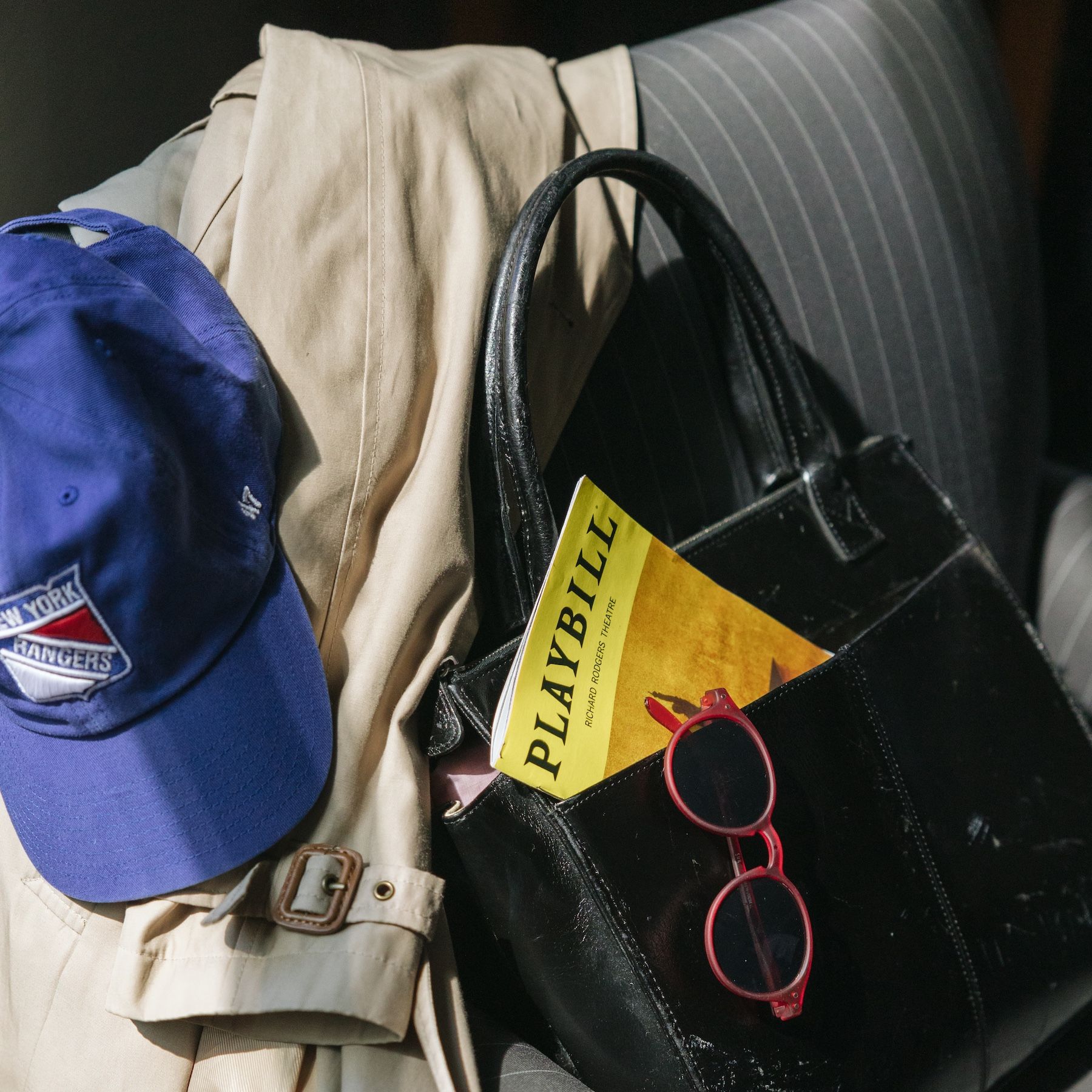 Blue baseball cap, black purse with sunglasses and Playbill, tan trench coat, pinstripe jacket.