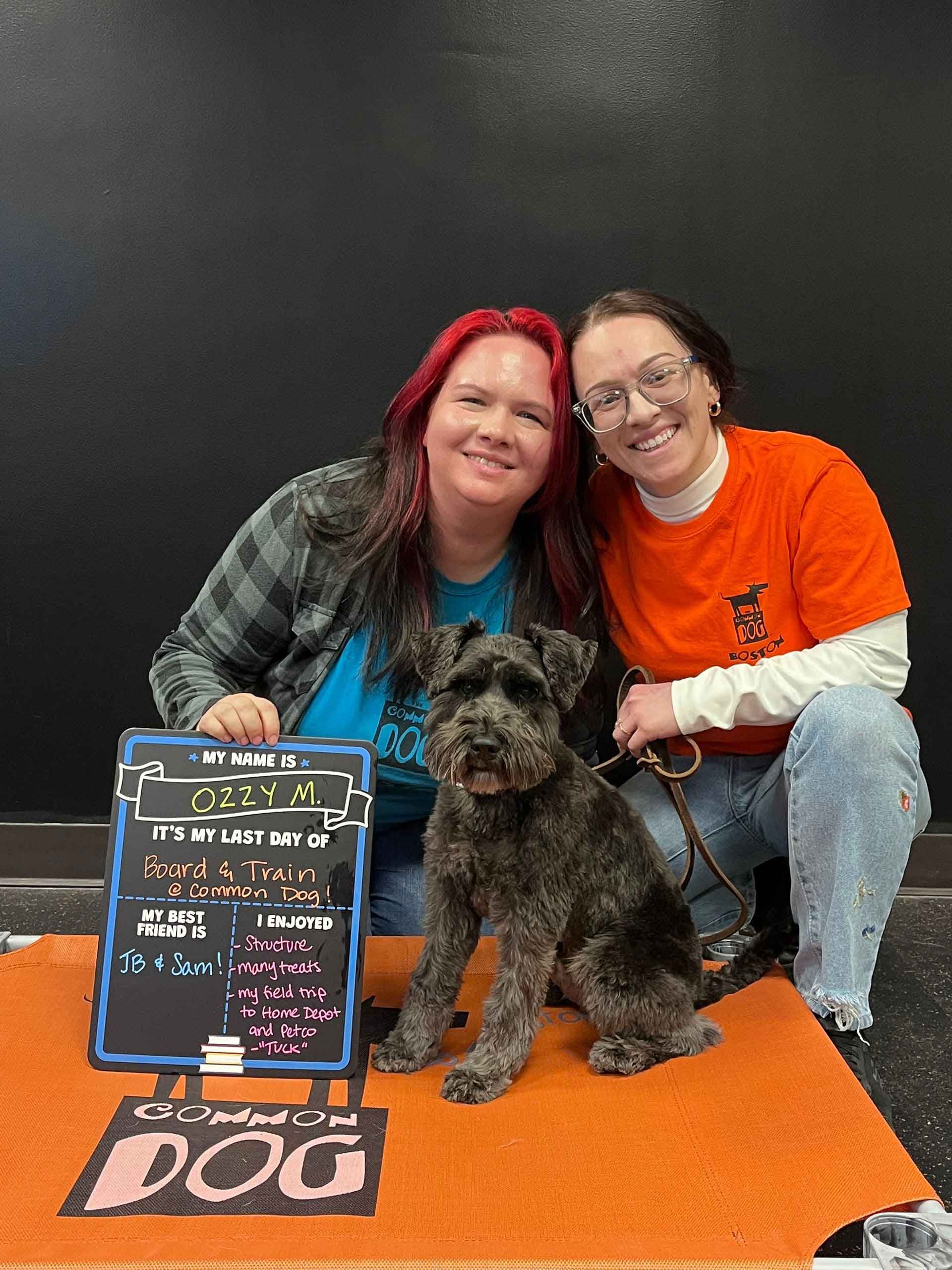 Two women are posing for a picture with a small black dog.