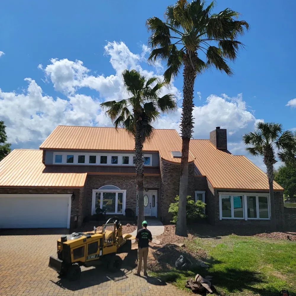 House with copper roof, palm trees, and worker with equipment in front.