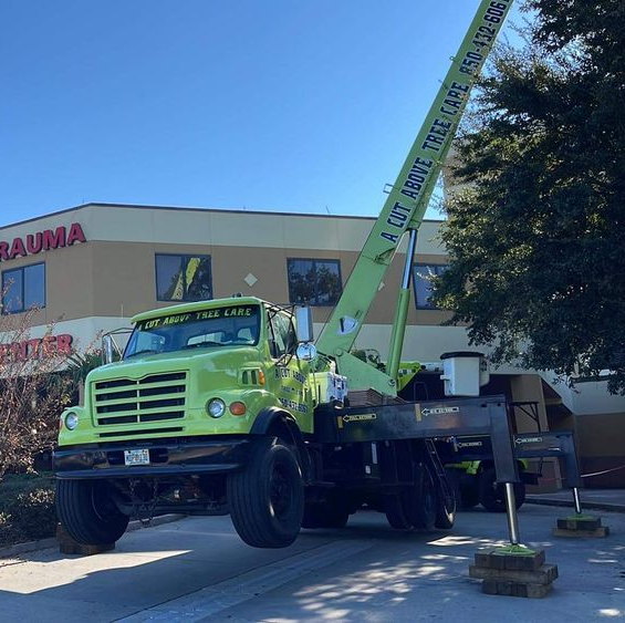 Green truck with crane in front of hospital emergency room; ambulance nearby under a clear blue sky.