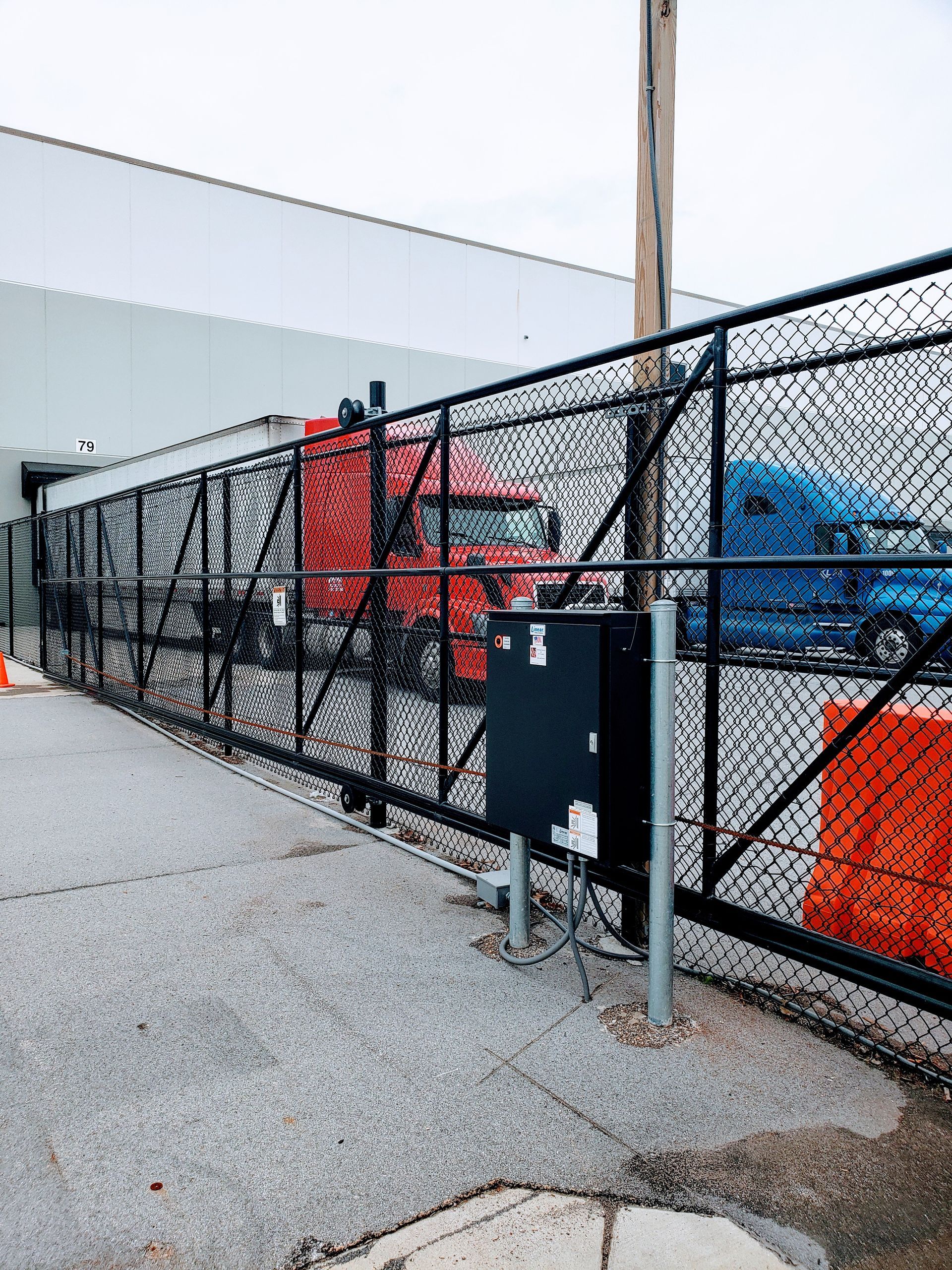 Black chain-link fence with sliding gate, control box, and parked semi-trucks.