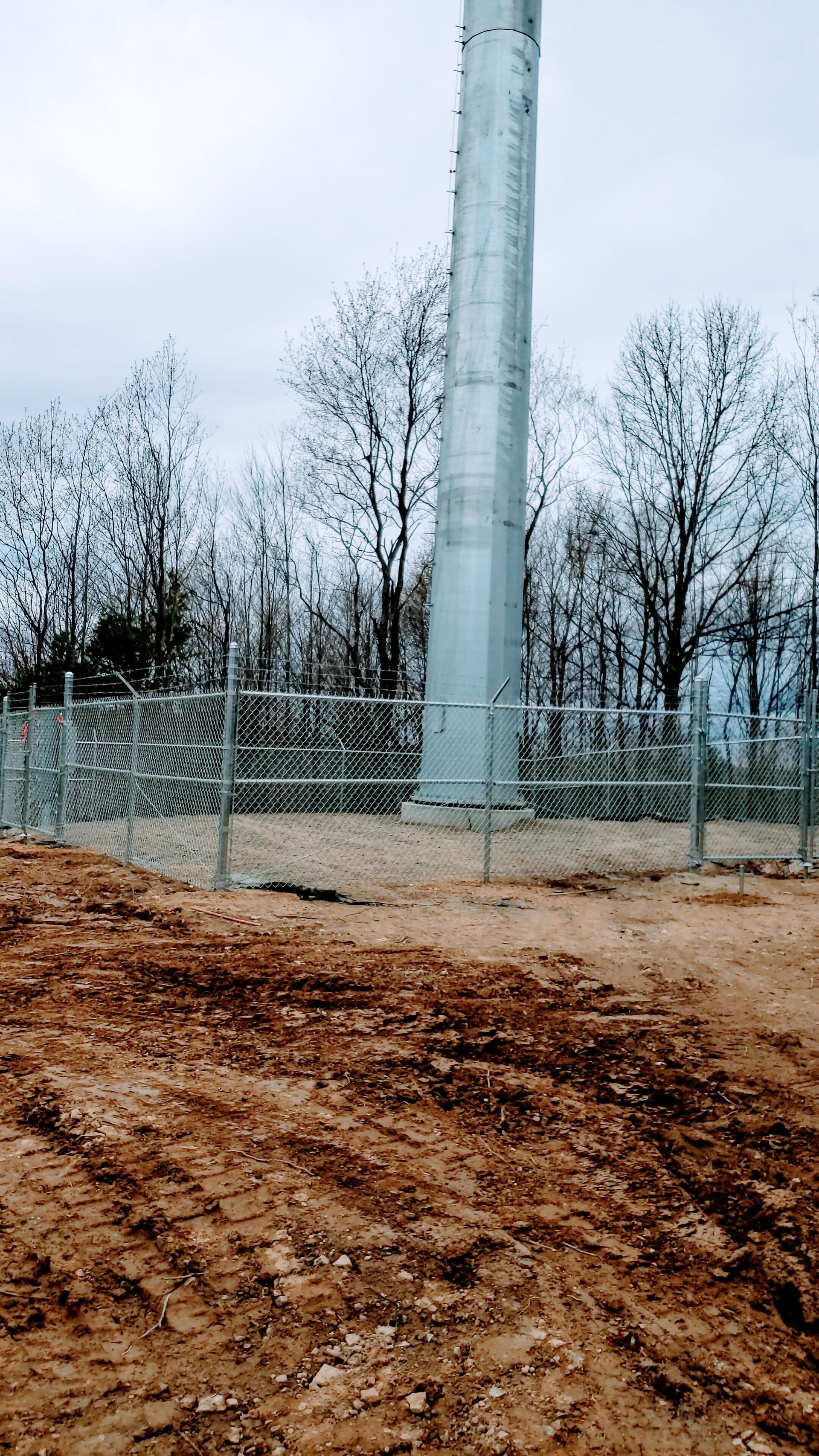 Cell tower surrounded by a chain link fence; brown dirt in the foreground and bare trees in the background on a cloudy day.