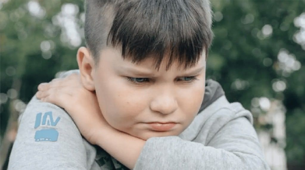 A young boy is sitting with his head resting on his arm and making a sad face.