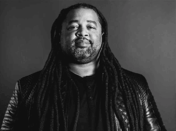 A black and white photo of a man with long dreadlocks and a beard.