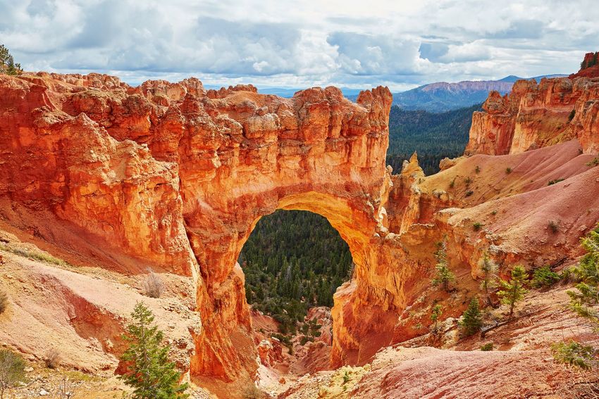 Red rock canyon with a natural stone arch under a cloudy sky, framed by rugged cliffs and sparse trees