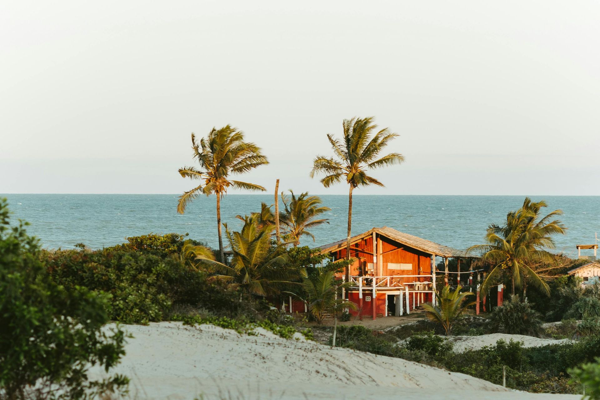 Red building with thatched roof, palm trees, and ocean view on a sandy beach.
