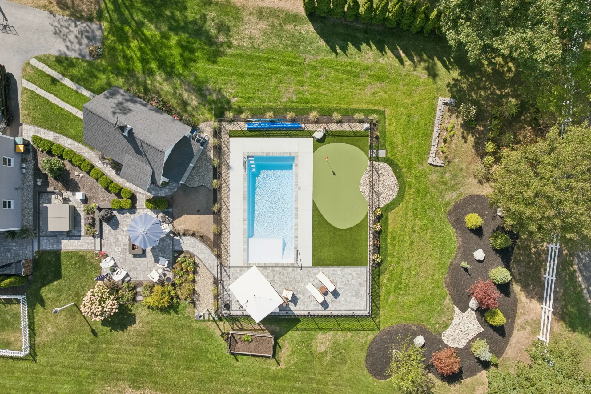 Overhead view of a backyard with a pool, putting green, patio, and landscaped areas on a green lawn.