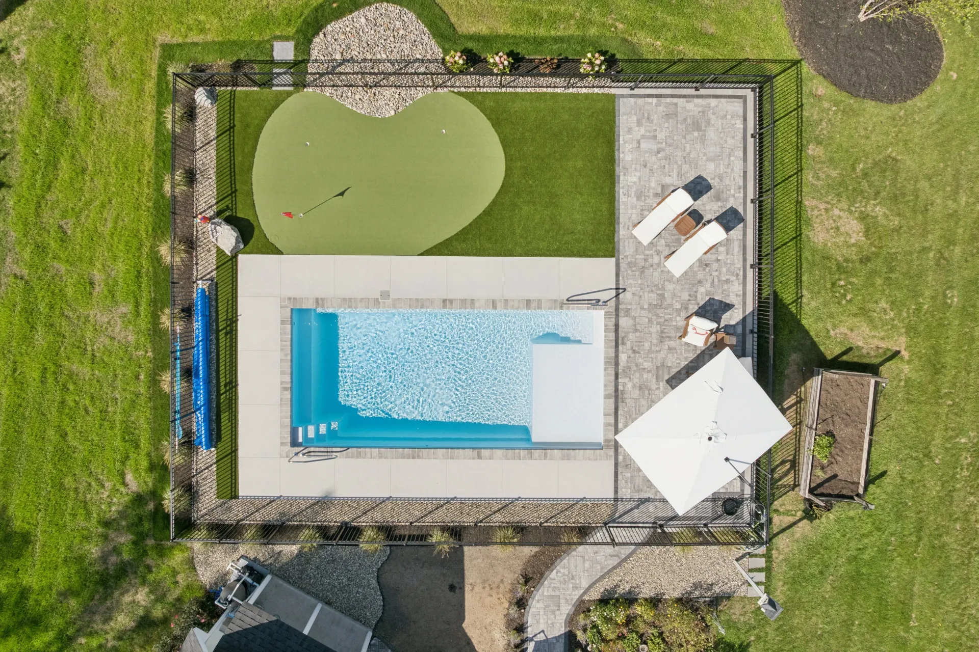 Overhead view of a rectangular pool, putting green, patio, and lawn. White chairs and umbrella are on the patio.