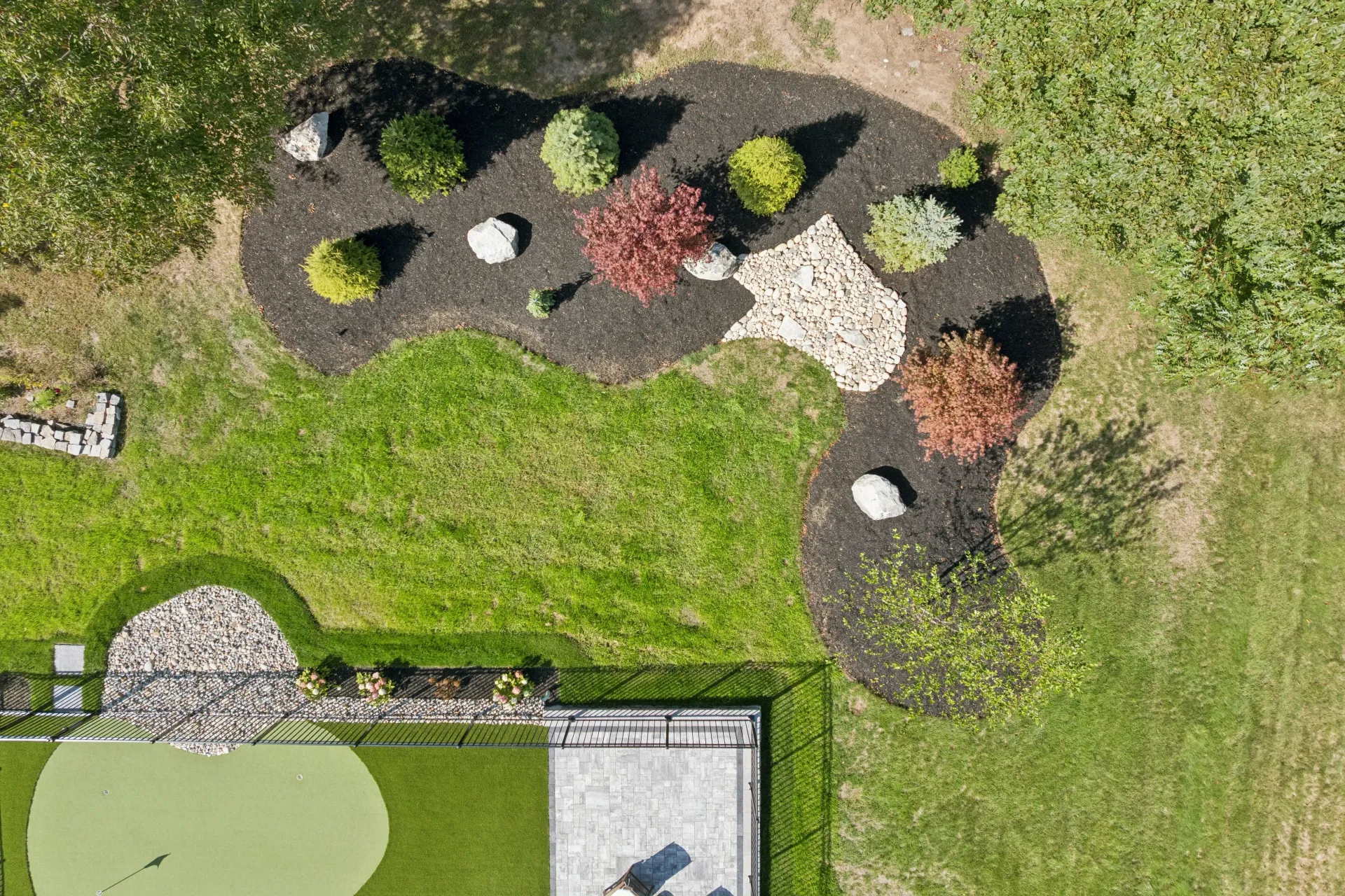 Overhead view of a landscaped area with mulch, plants, rocks, and a putting green surrounded by green grass.
