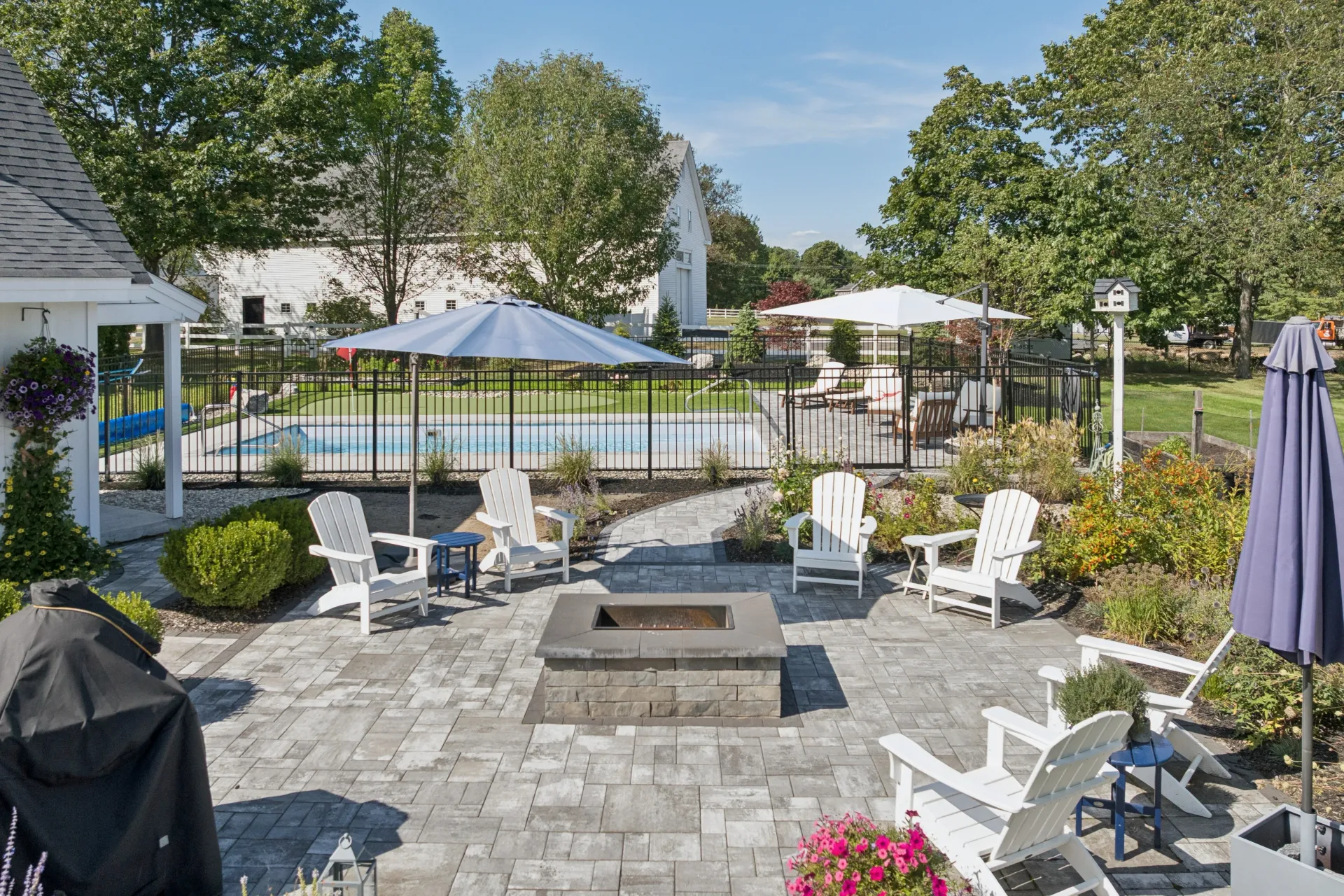 Backyard patio with fire pit, white Adirondack chairs, and a pool. Blue umbrellas and green foliage.