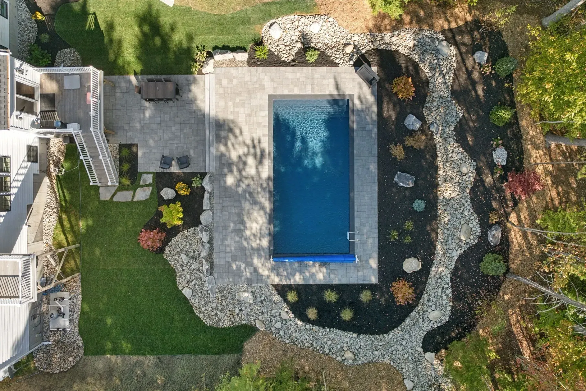 Aerial view of a rectangular pool surrounded by a stone patio, landscaping, and lush green lawn.