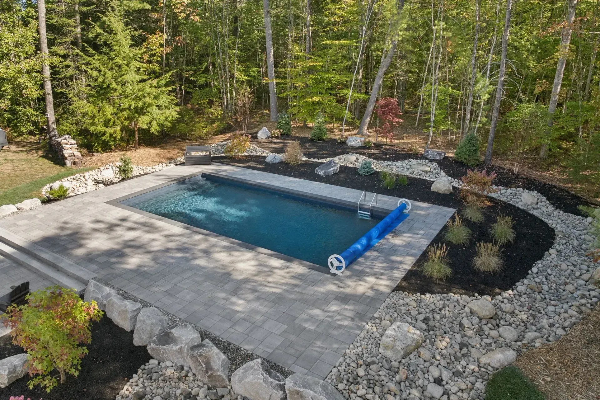 Rectangular in-ground pool surrounded by rocks, greenery, and trees. Blue pool cover is partially rolled up.