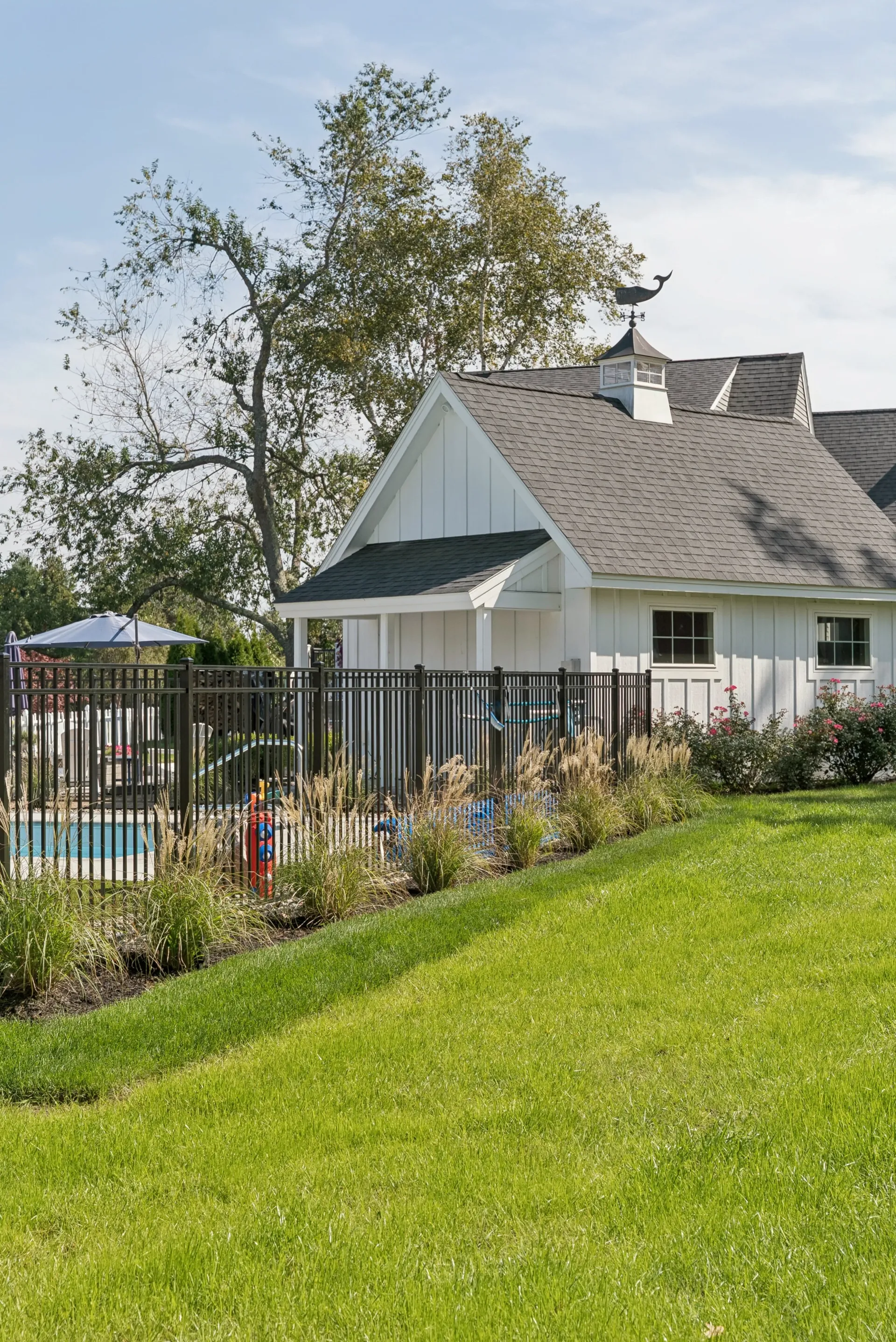 White building with dark roof near a pool, framed by a black fence and a grassy hill.