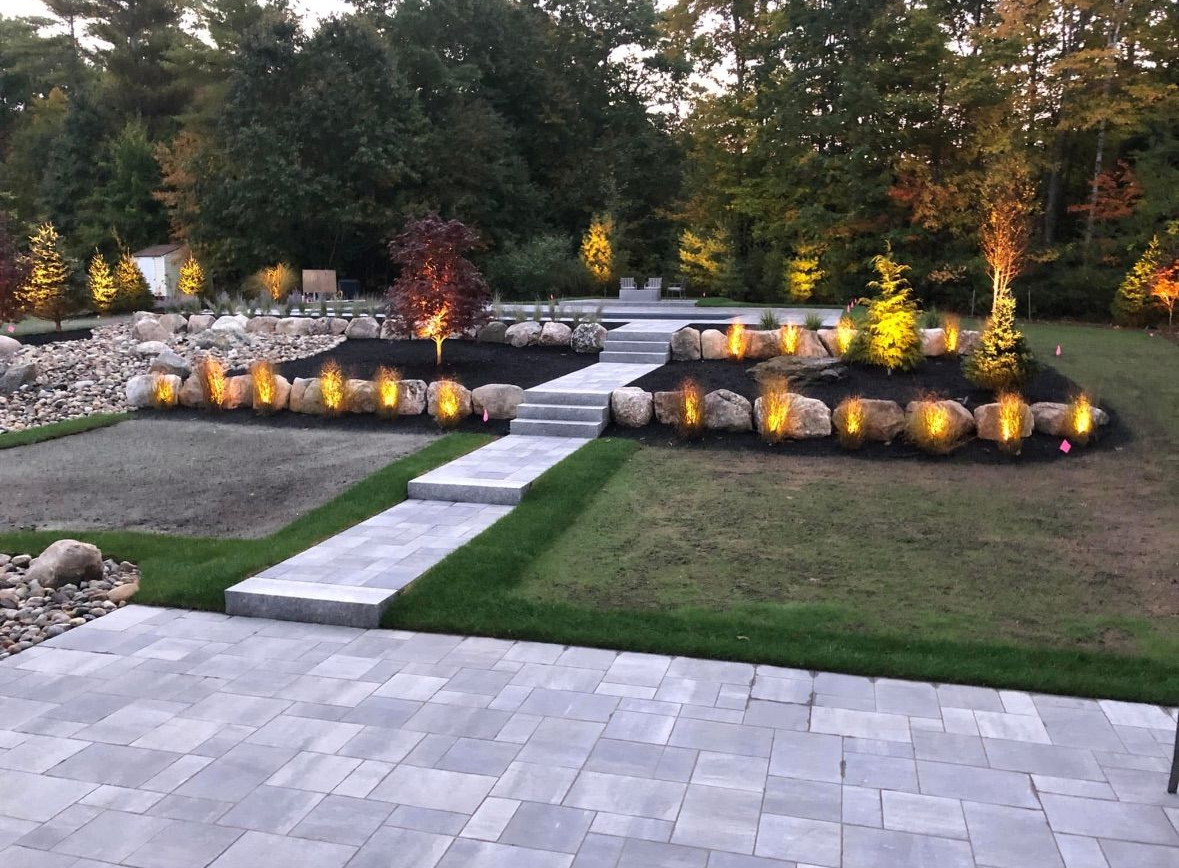 Backyard with illuminated landscaping, stone steps, lawn, and patio at dusk.