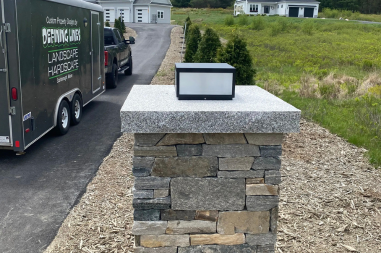 Stone pillar with a granite top and a modern light, set in front of a driveway and grass.