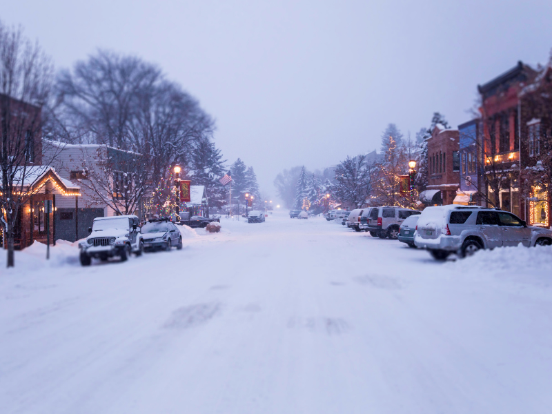 Snowy street lined with buildings and parked cars; overcast sky.