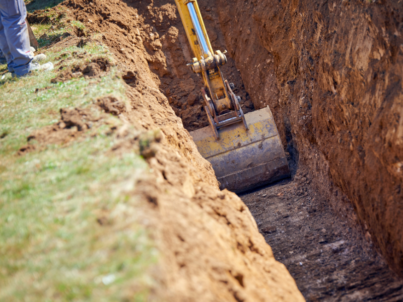 Excavator digging a trench in the earth, removing soil, with a person's legs visible nearby.