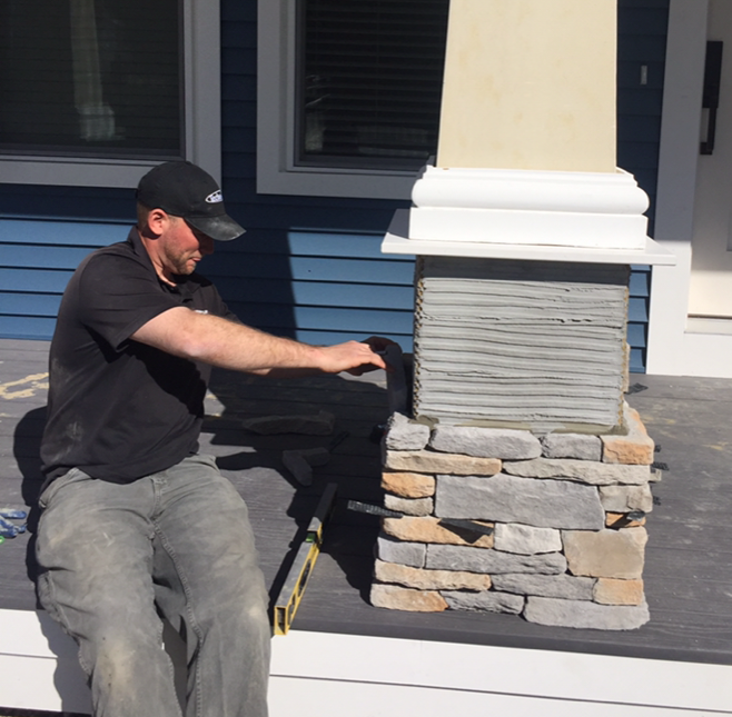 Man installing stone veneer on a porch column, wearing a cap and jeans, blue siding in background.