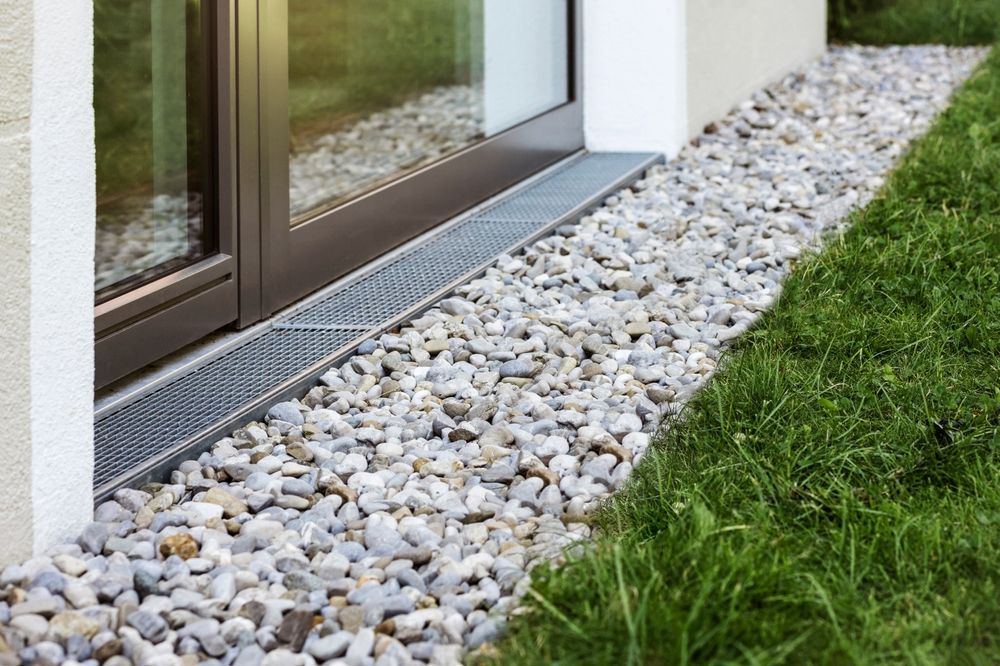 Gravel bed with a metal drain next to a glass window and a green lawn.
