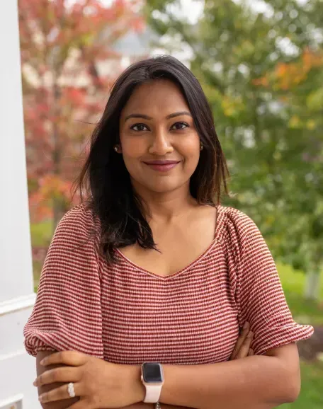 Woman with brown skin, arms crossed, wearing a pink striped shirt, smiling outside with trees in the background.