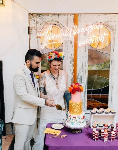 A bride and groom cut a colorful wedding cake. The bride wears a floral crown, and a neon sign reads